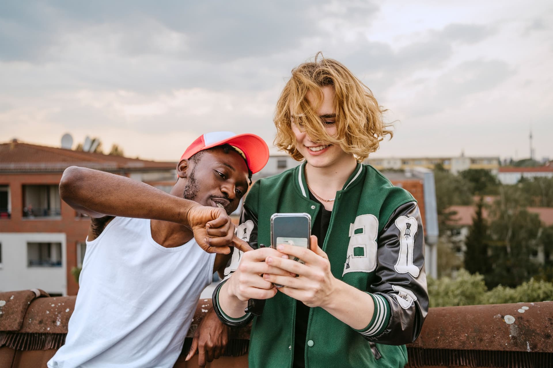 Zwei junge, erwachsene Männer, in sportlich modischer, Freizeitkleidung, stehen auf dem Dach in einer Wohnsiedlung und sehen zusammen auf den Screen eines Smartphones. 