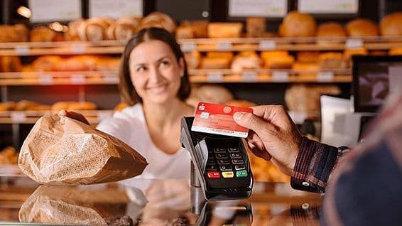 Person hält Sparkassenkarte an ein Kartenlesegerät in einer Bäckerei während Verkäuferin eine Brottüte überreicht