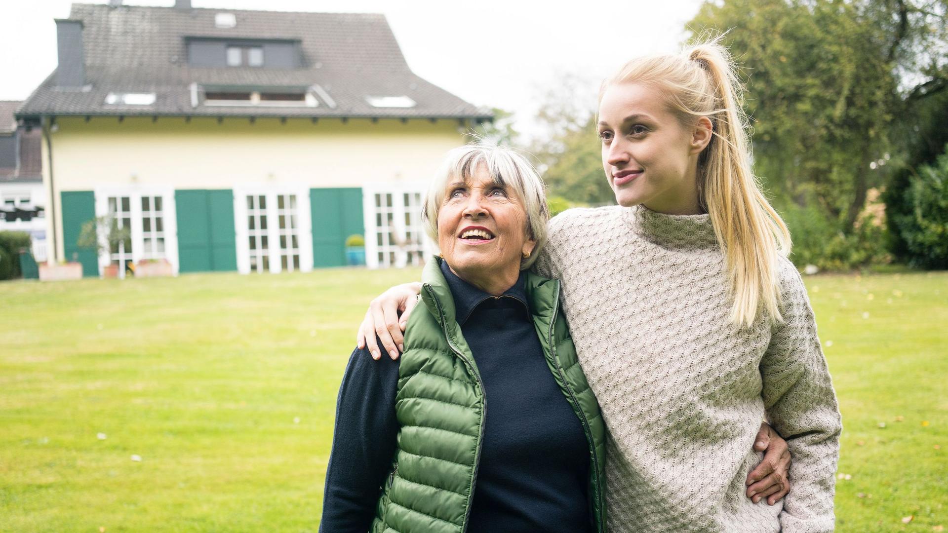 Eine junge Frau mit langem blonden Pferdeschwanz legt ihren Arm um ihre Großmutter. Sie stehen auf der Wiese eines Grundstücks. Im Hintergrund steht ein schönes altes Wohnhaus. Beide lächeln.