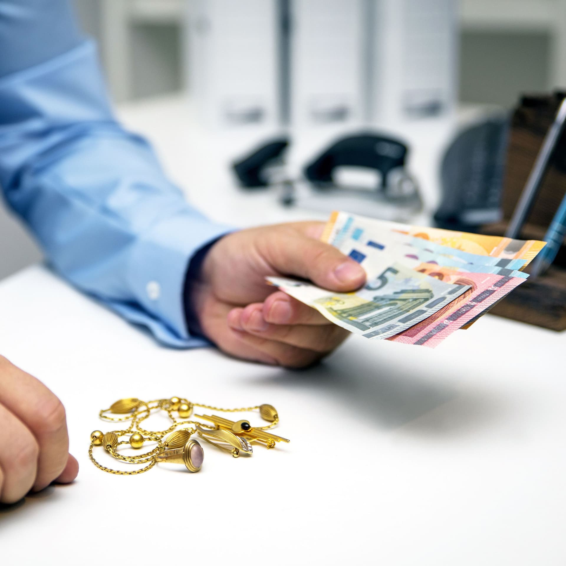 Auf einem Tisch liegt Goldschmuck. Eine Hand hält Bargeld. Szene in einem Pfandleihhaus.