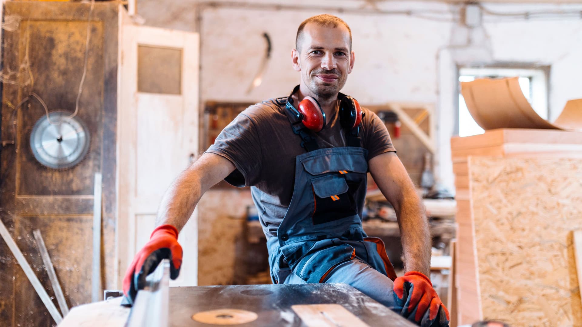 Mann mit Arbeitshandschuhen lehnt in einer Schreinerei an einer Werkbank und lächelt in die Kamera.