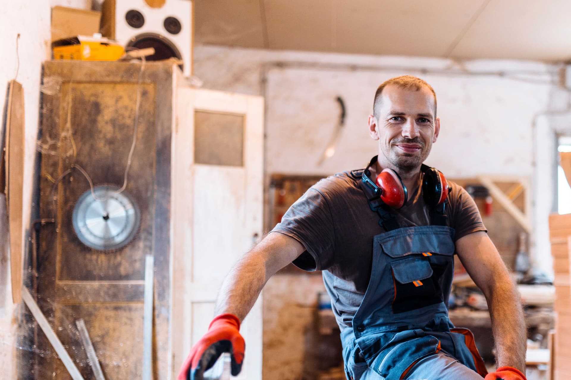 Mann mit Arbeitshandschuhen lehnt in einer Schreinerei an einer Werkbank und lächelt in die Kamera.