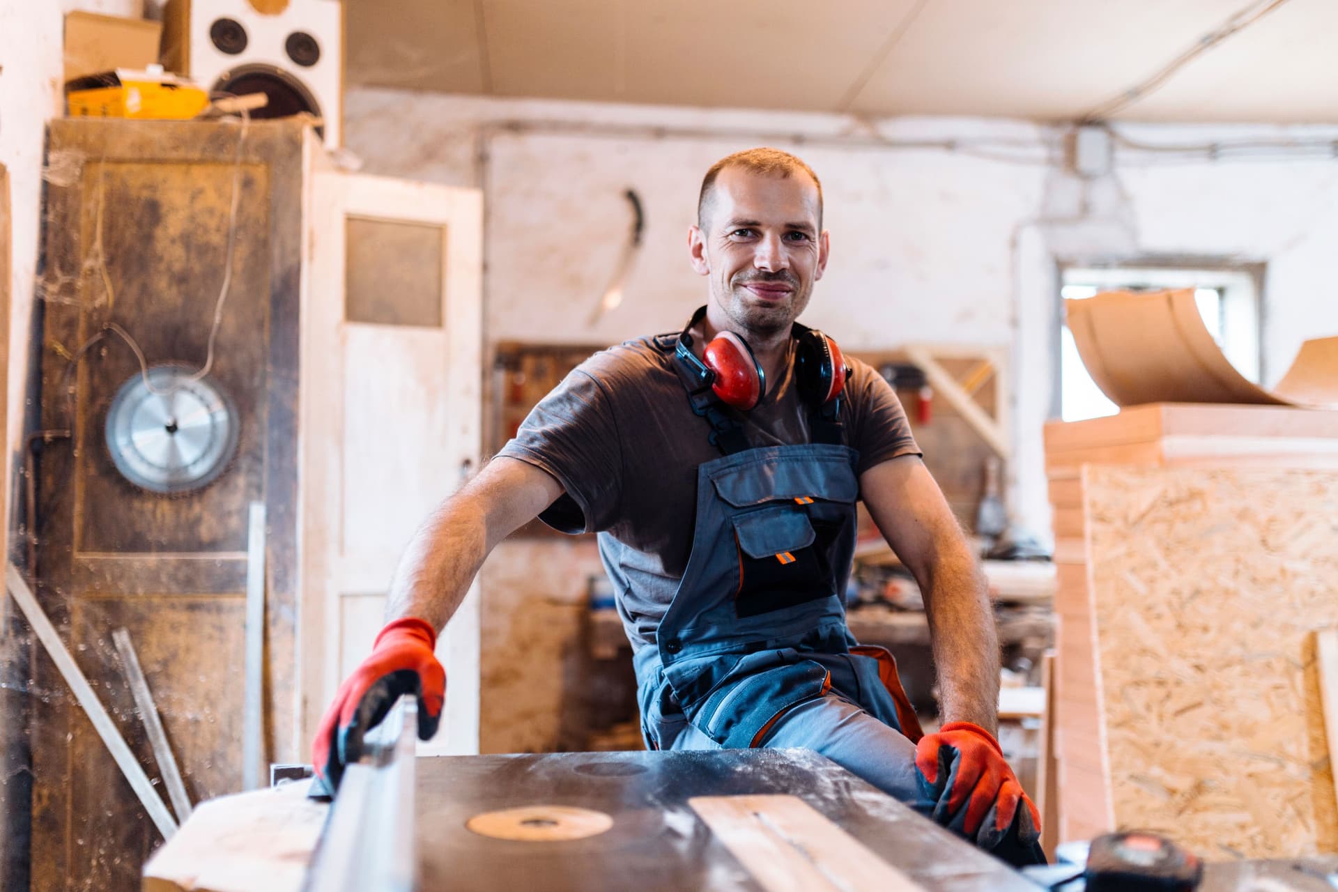Mann mit Arbeitshandschuhen lehnt in einer Schreinerei an einer Werkbank und lächelt in die Kamera.