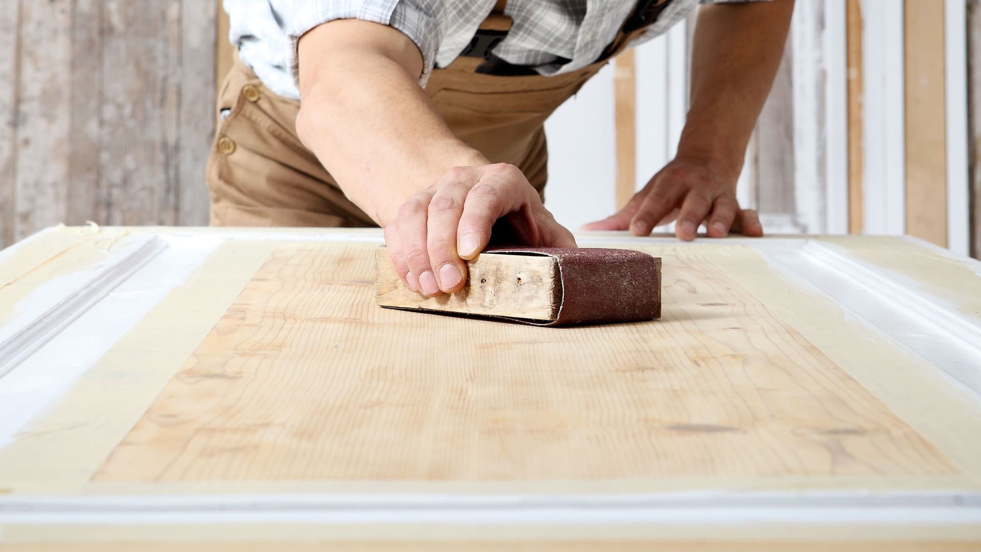 Schreiner mit Overall und Mütze, der in einer Schreinerei Holz bearbeitet und eine Holztür mit Schleifpapier abschleift.