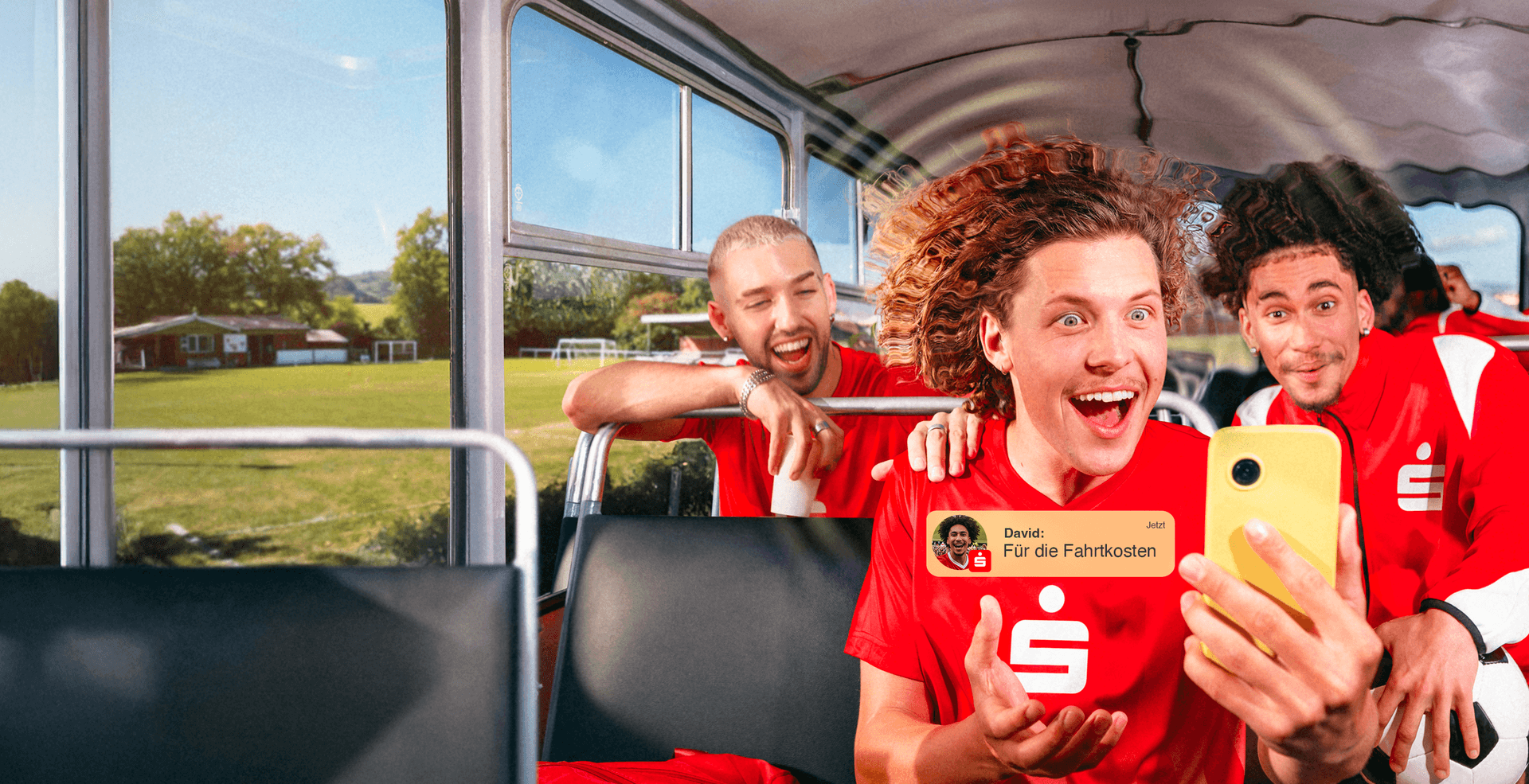 Junge Fußballspieler in roten Trikots sitzen fröhlich in einem Bus; im Vordergrund hält ein Spieler ein gelbes Smartphone, im Bild ist zusätzlich eine Push-Nachricht zu sehen: „David: Für die Fahrtkosten“.