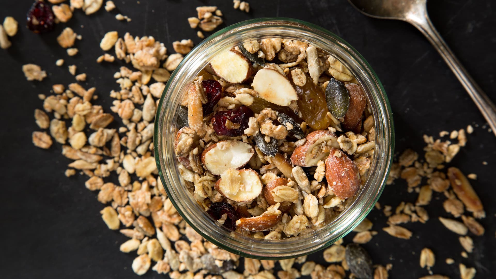 Müsli mit Nüssen in einem Glas und neben dem Glas auf einem schwarzen Untergrund verstreut. Daneben liegt ein Löffel.