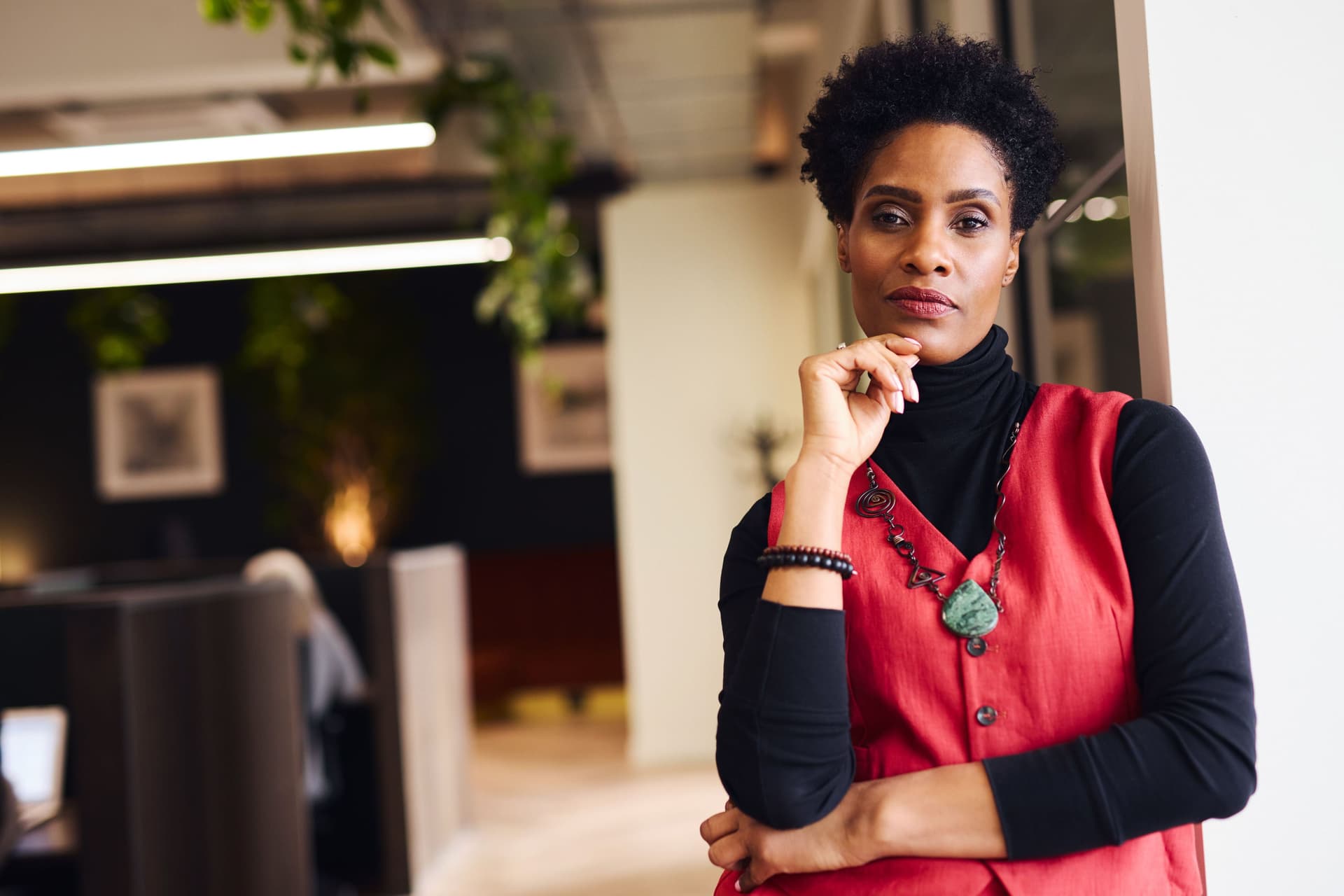 Porträt einer mittelalten Frau mit der Hand am Kinn, die mit nachdenklicher Miene im Coworking Space in die Kamera blickt.