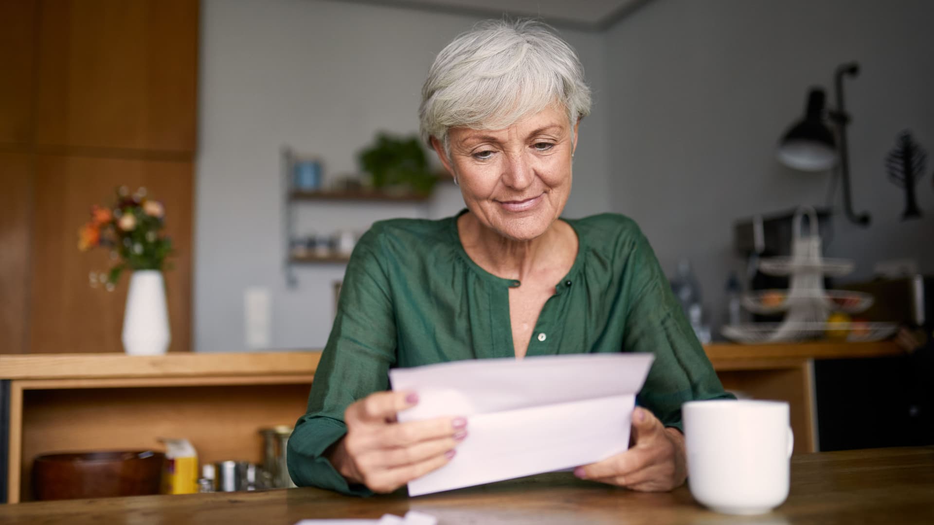 Seniorin sitzt mit Kaffeetasse an einem Tisch und liest lächelnd einen Brief.