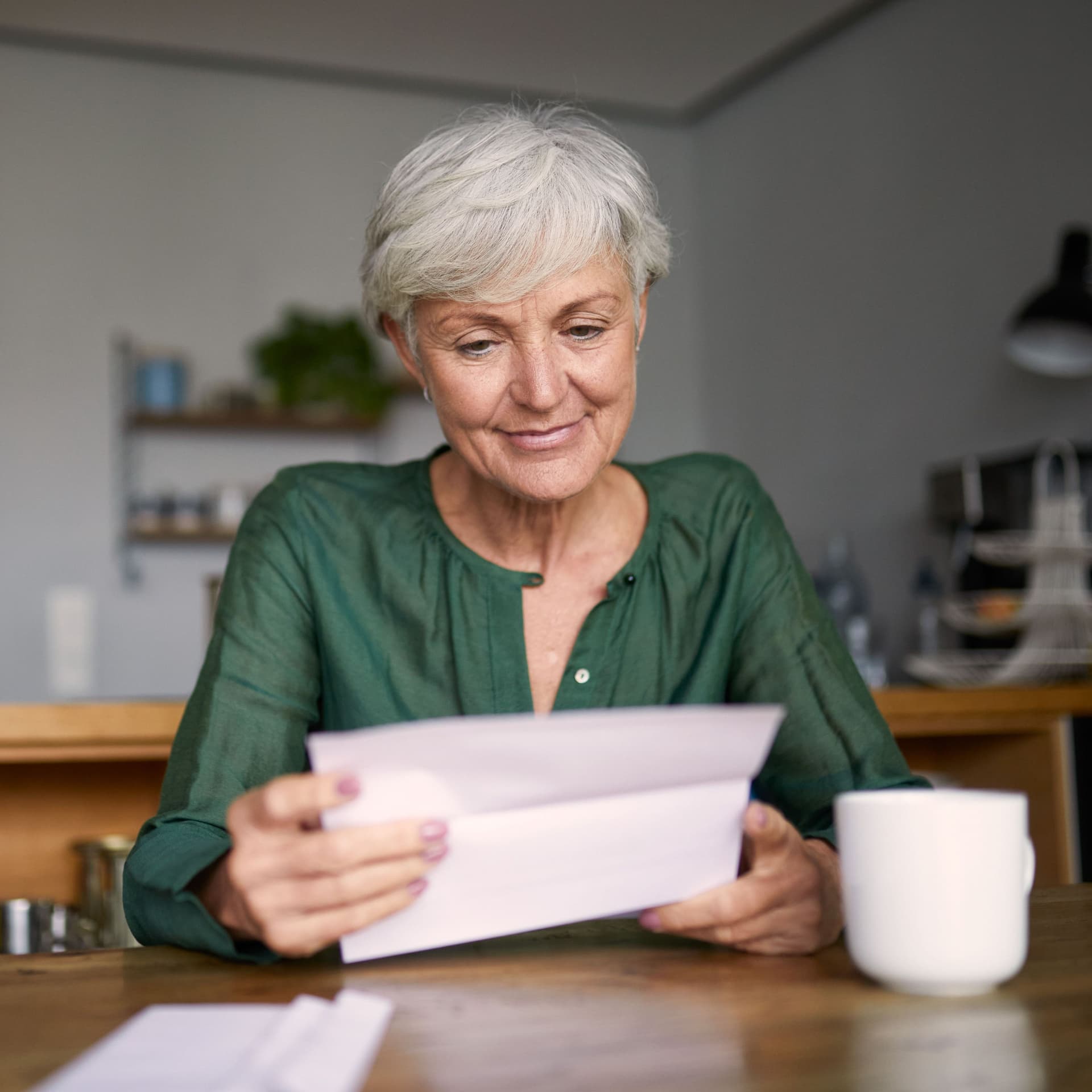 Seniorin sitzt mit Kaffeetasse an einem Tisch und liest lächelnd einen Brief.