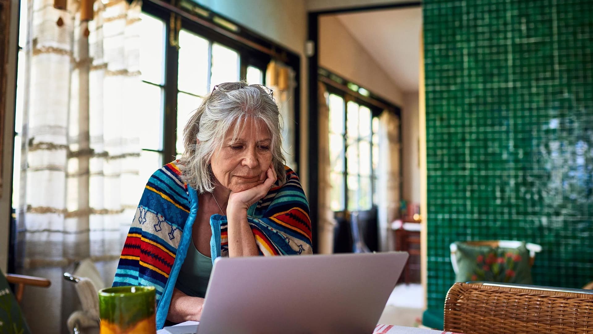 Eine Seniorin sitzt nachdenklich vor einem Laptop an einem Tisch in einem gemütlichen, farbenfrohen Raum. Sie trägt eine Boho-Decke über ihren Schultern.