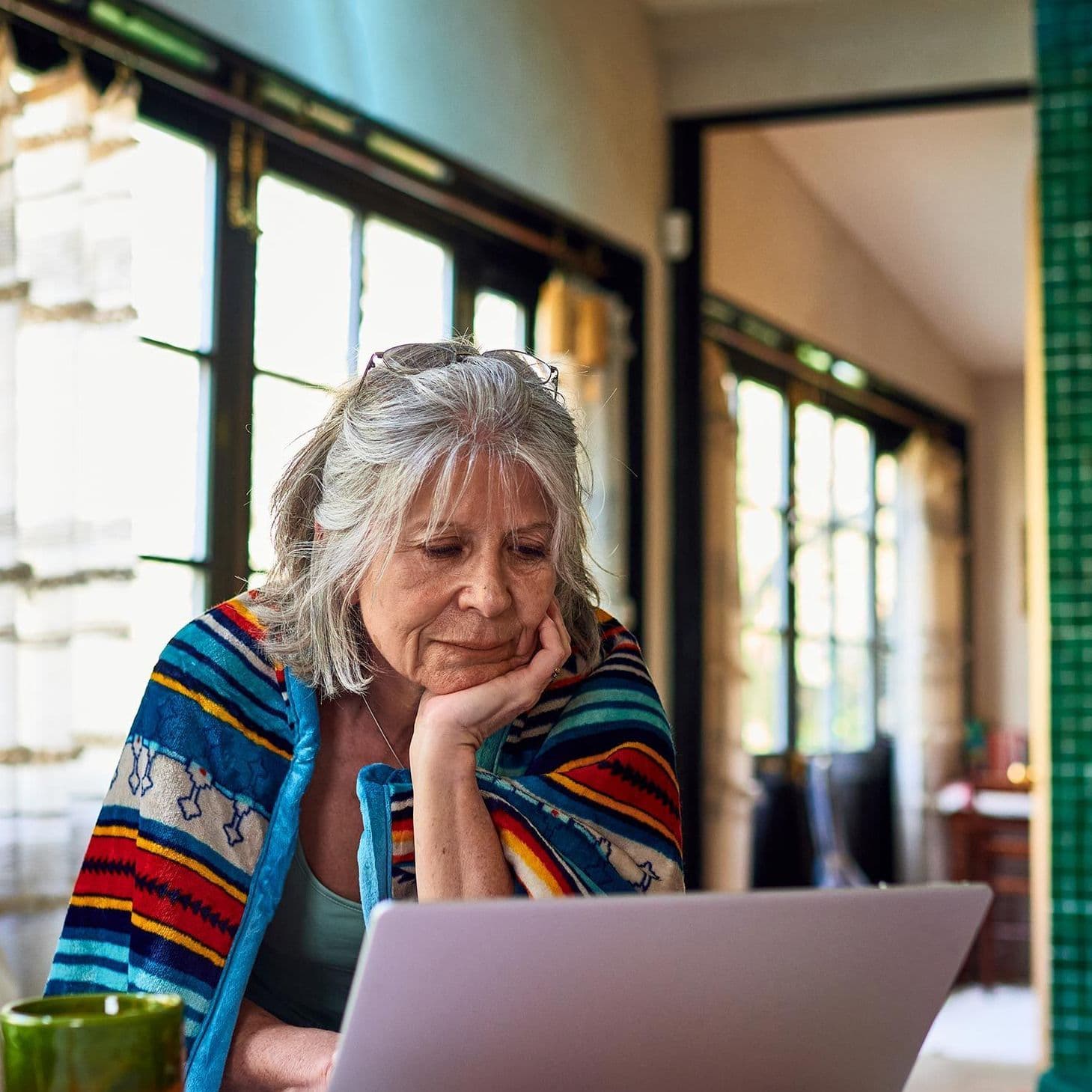 Eine Seniorin sitzt nachdenklich vor einem Laptop an einem Tisch in einem gemütlichen, farbenfrohen Raum. Sie trägt eine Boho-Decke über ihren Schultern.