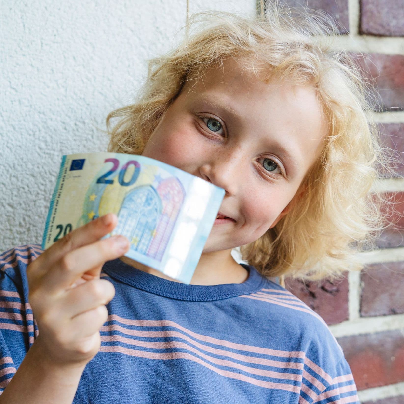 Eine bunte Bildkomposition mit einem Jungen im Grundschulalter, der freudig einen Zwanzig-Euro-Schein in die Kamera hält.