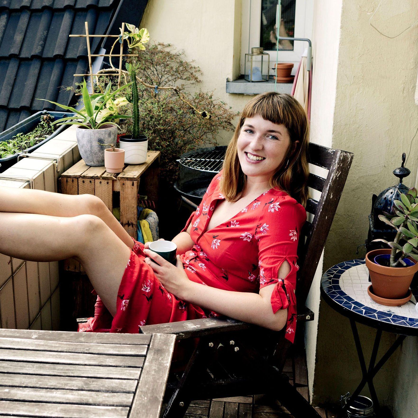 Frau in rotem Kleid sitzt auf einem Balkon mit den Füßen auf der Balkonbrüstung, es ist Frühling