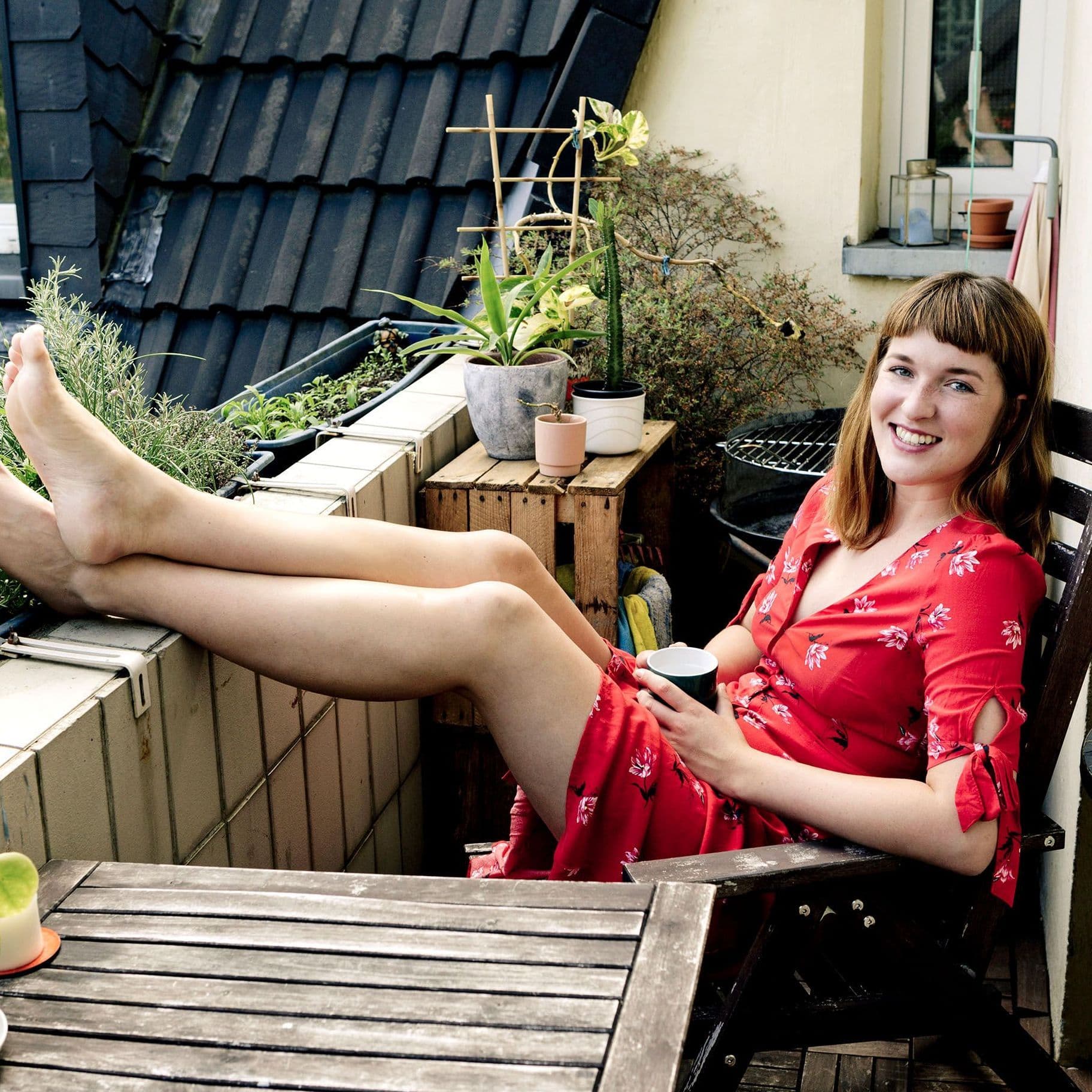 Frau in rotem Kleid sitzt auf einem Balkon mit den Füßen auf der Balkonbrüstung, es ist Frühling