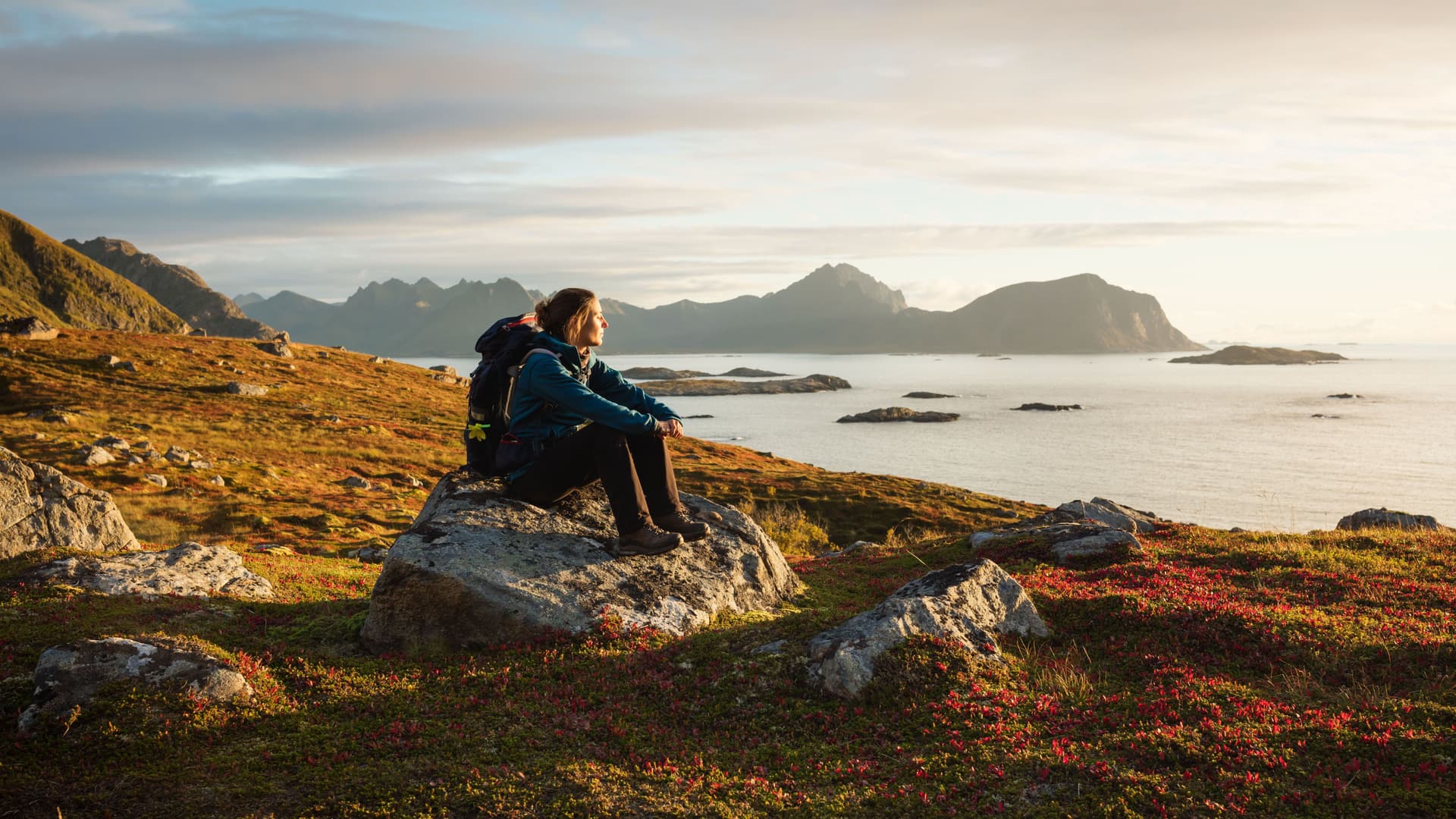Eine Frau sitzt in einer schönen Landschaft in Wanderbekleidung auf einem Stein und schaut in die Ferne.