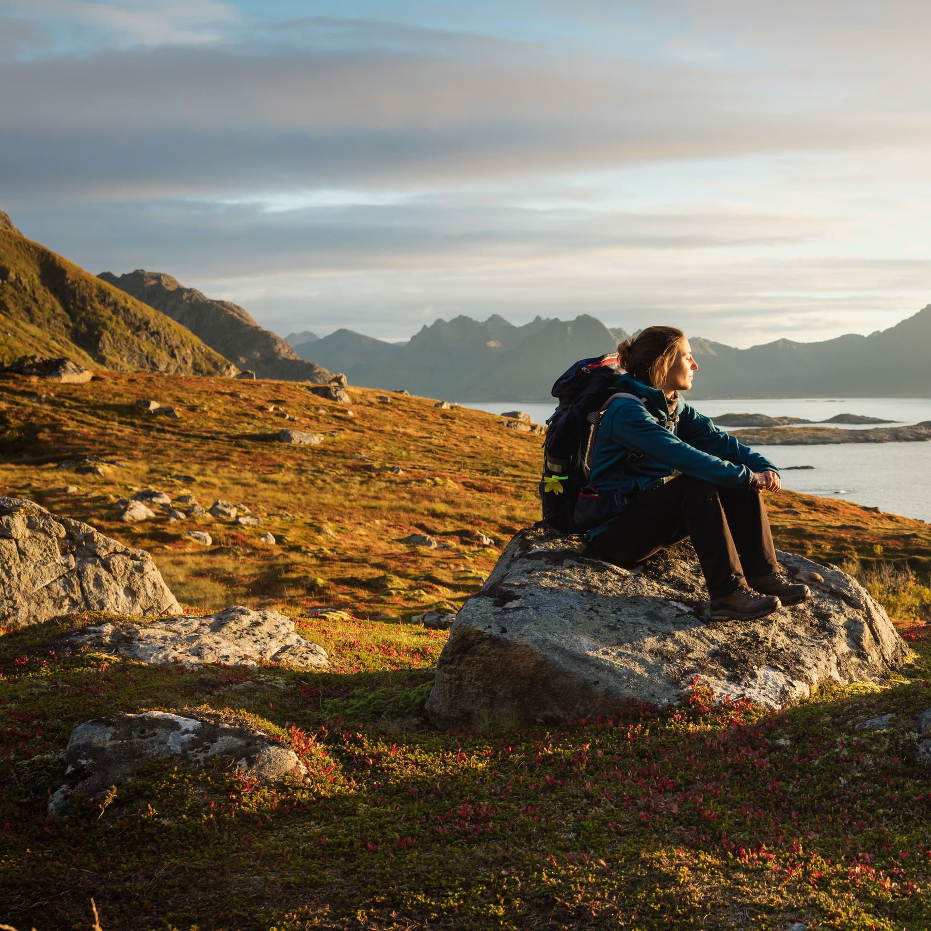 Eine Frau sitzt in einer schönen Landschaft in Wanderbekleidung auf einem Stein und schaut in die Ferne.