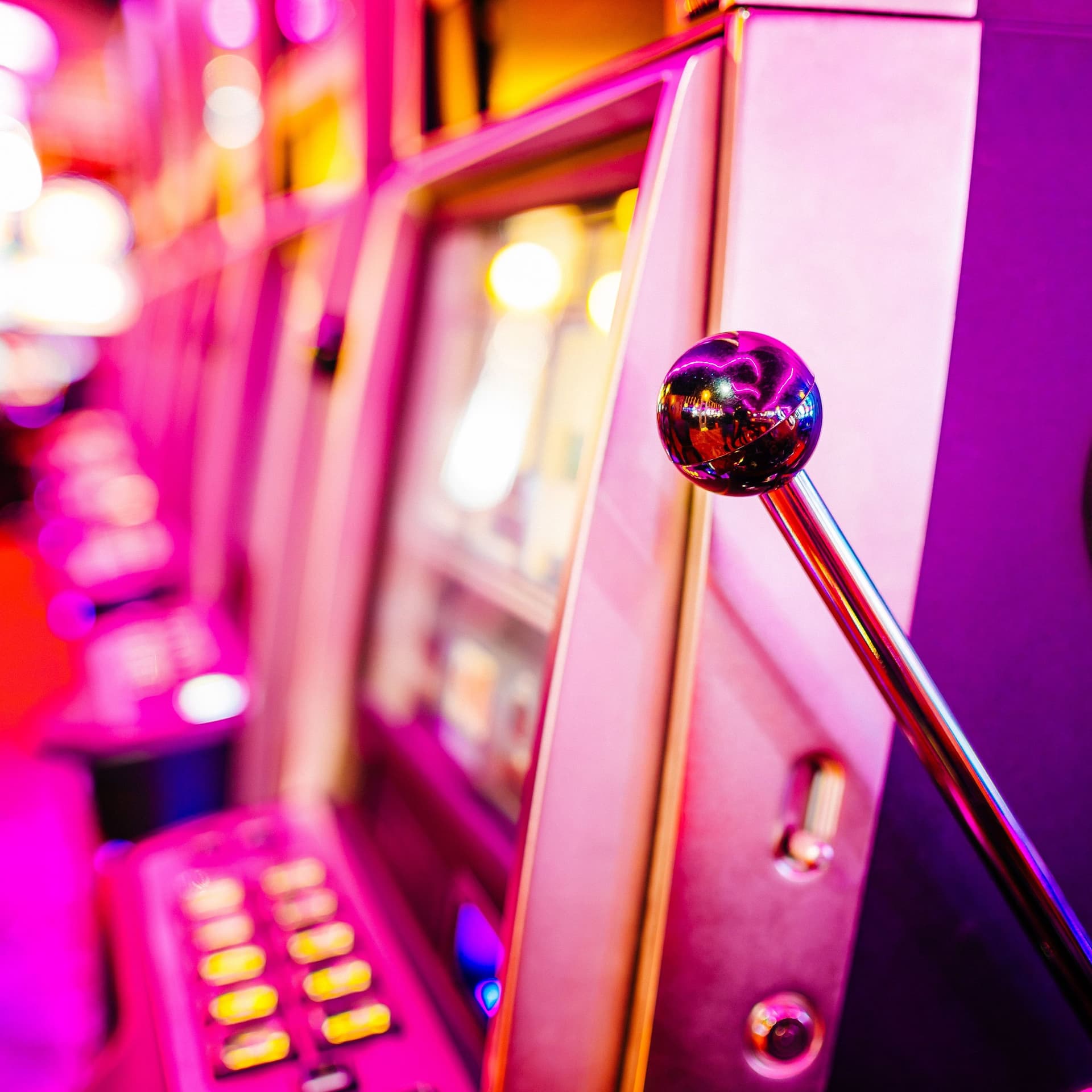 Seitliche Aufnahme auf eine Reihe von Spielautomaten mit Sitzplätzen davor. Sie werden von buntem LED Licht angestrahlt.
