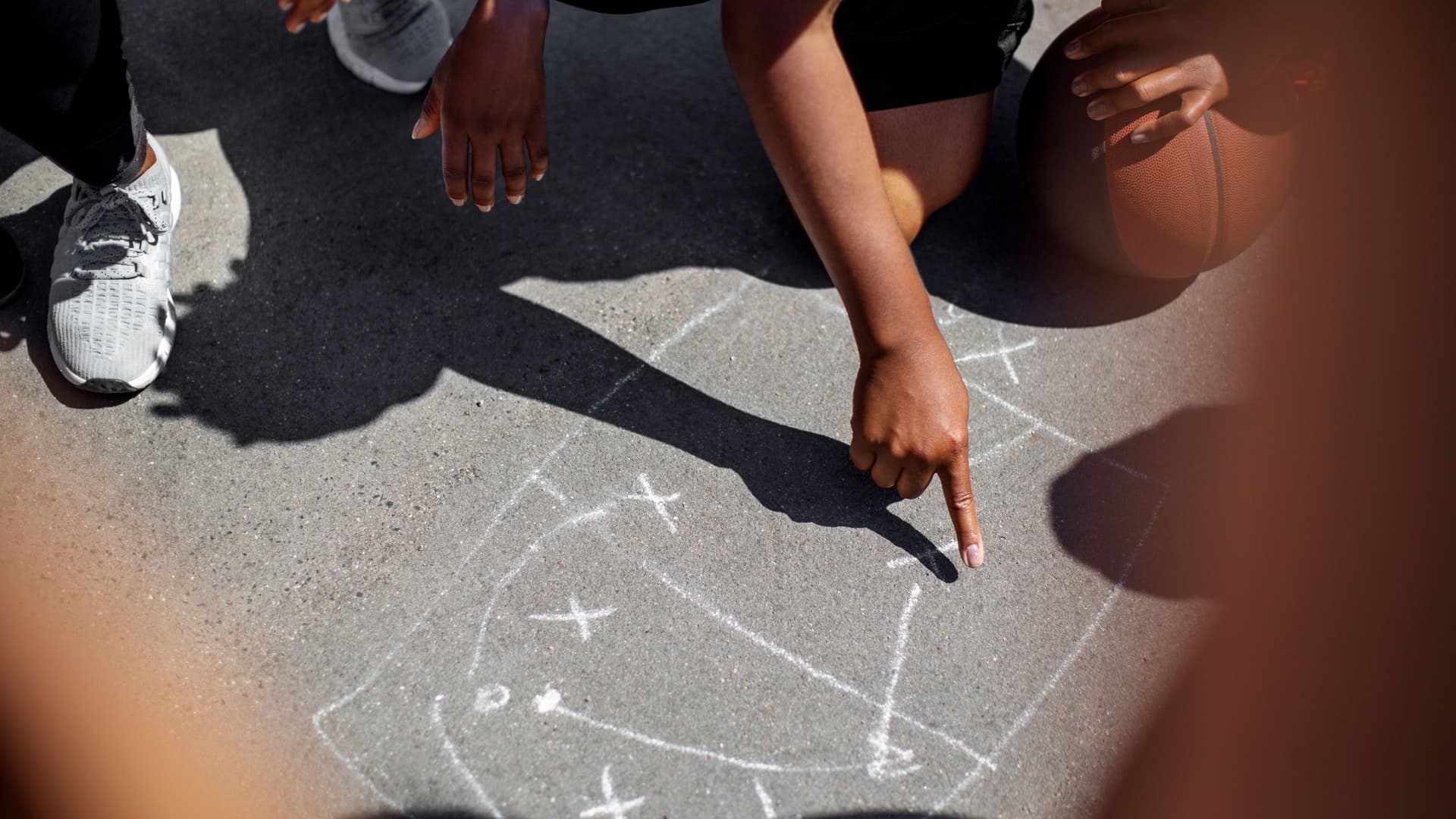 Nahaufname von BPOC Basketballspielern die Spielzüge mit Kreide auf den Boden gemalt haben. Zu sehen sind nur Hände und Knie.