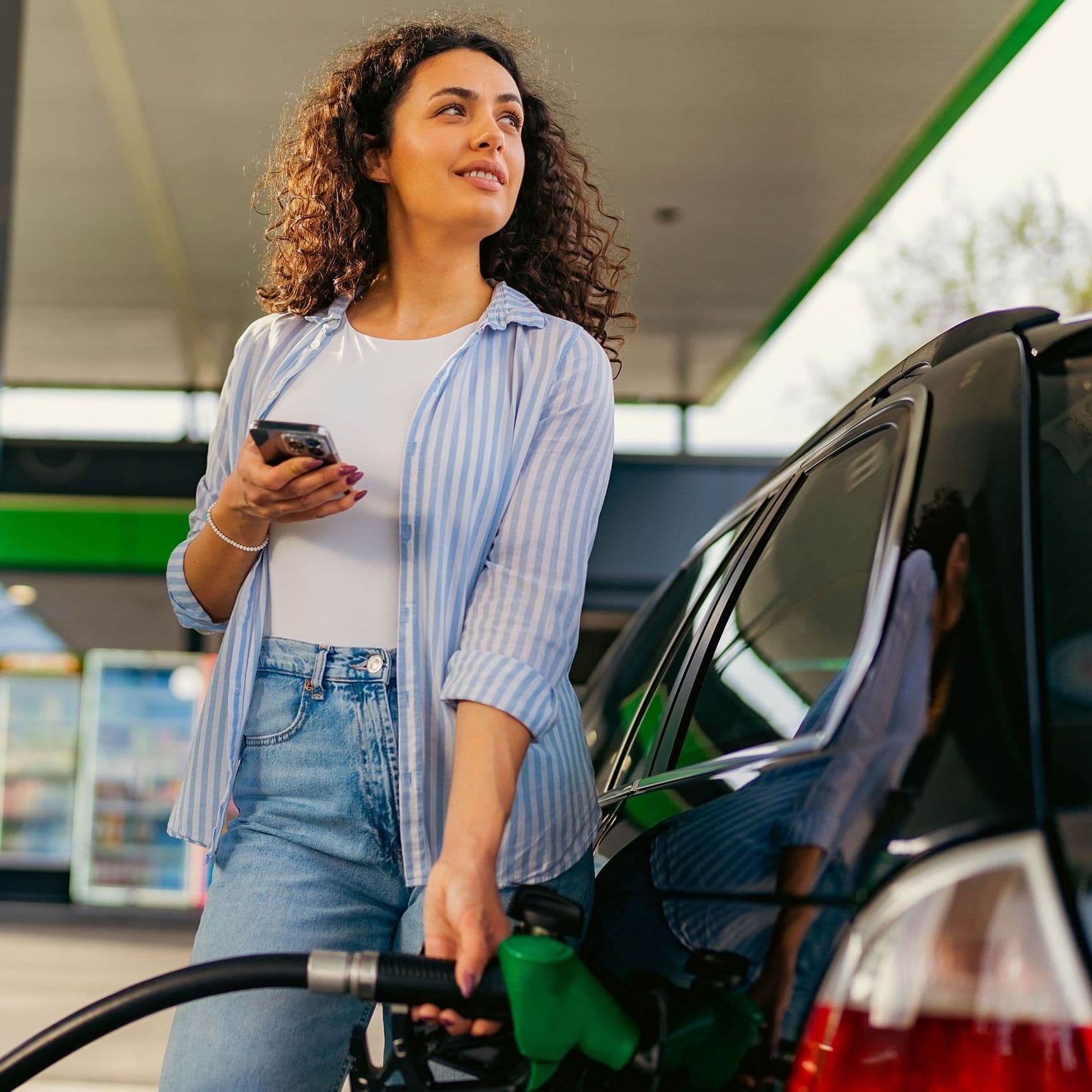 Eine Frau mit lockigen Haaren und sommerlicher Kleidung tankt an einer Tankstellenzapfsäule Benzin. Sie hält ein Smartphone in der Hand und schaut nach oben auf die Preistafel. 