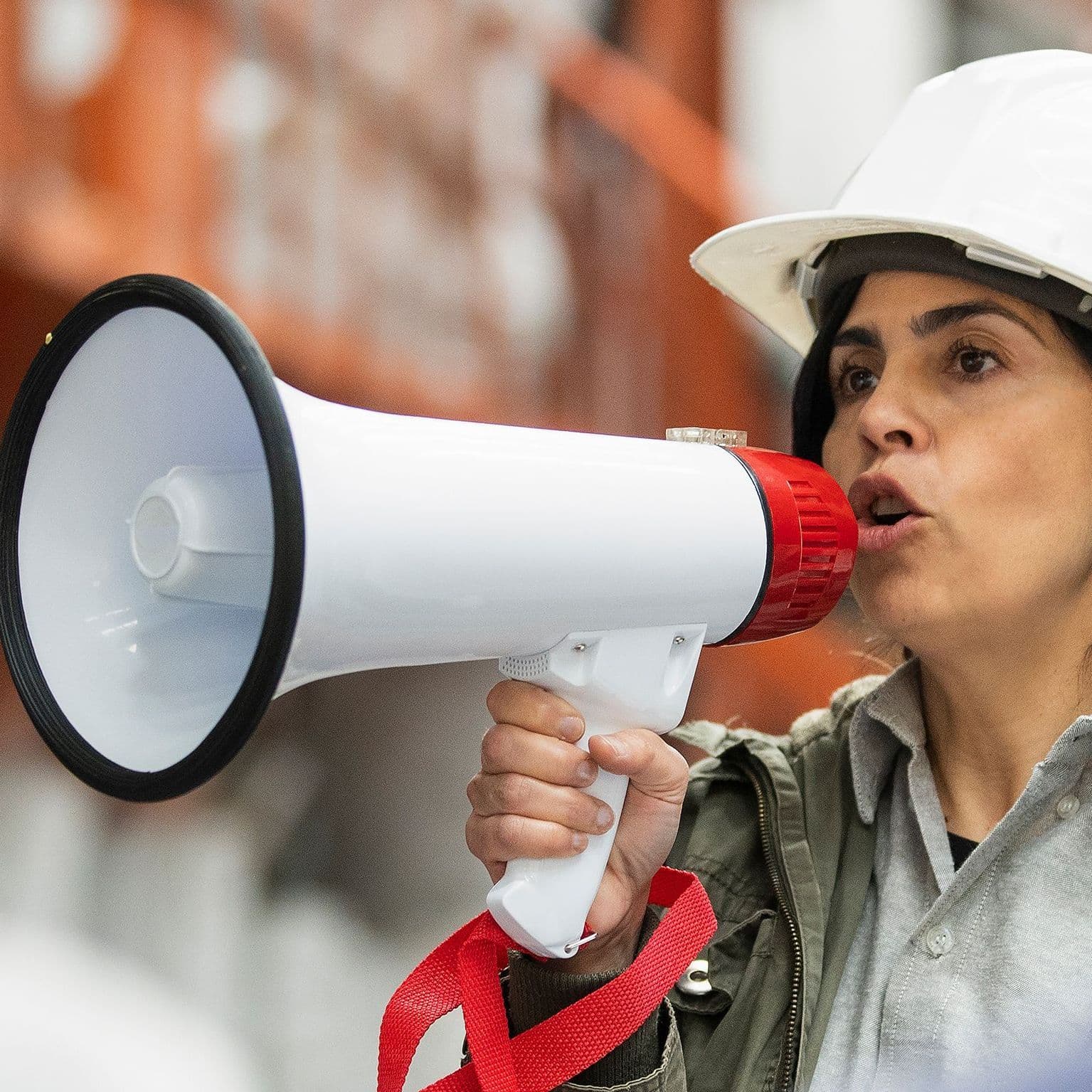 Eine Frau mit Bauhelm spricht auf einem Firmengelände vor in ein Megaphon.