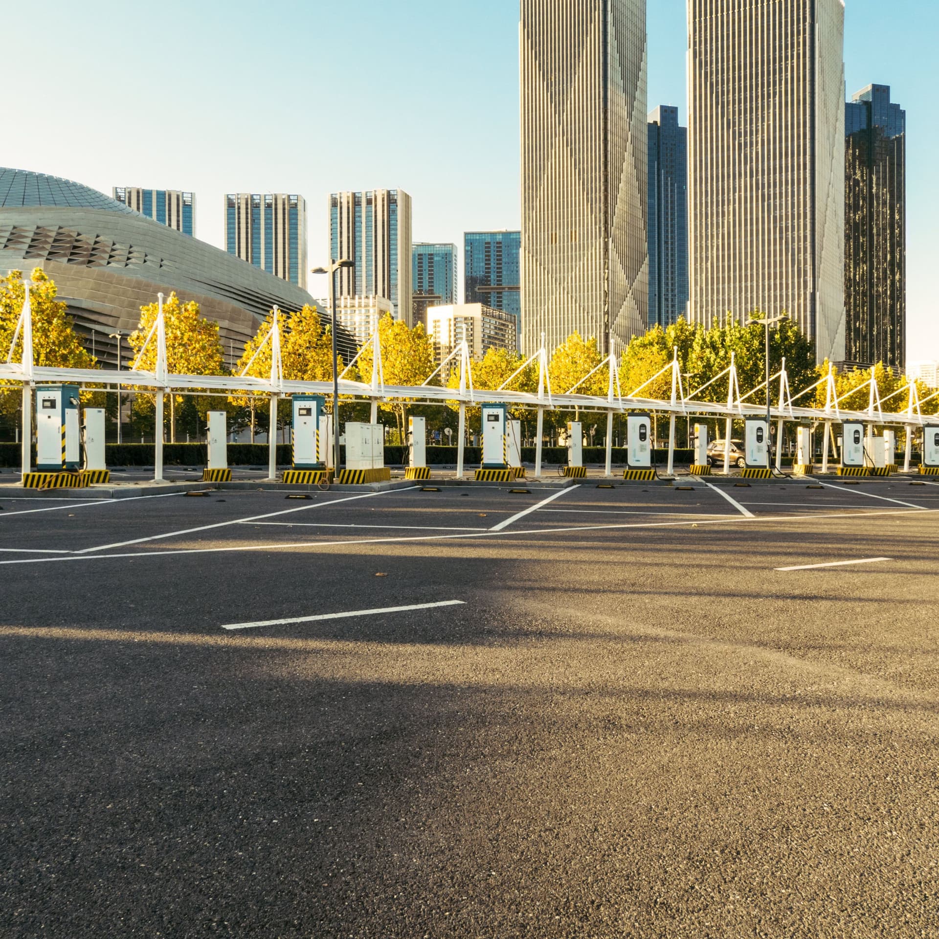 Ein großer leerer Parkplatz mit Aufladestationen für Elektroautos. Im Hintergrund Hochhäuser im Sonnenschein.