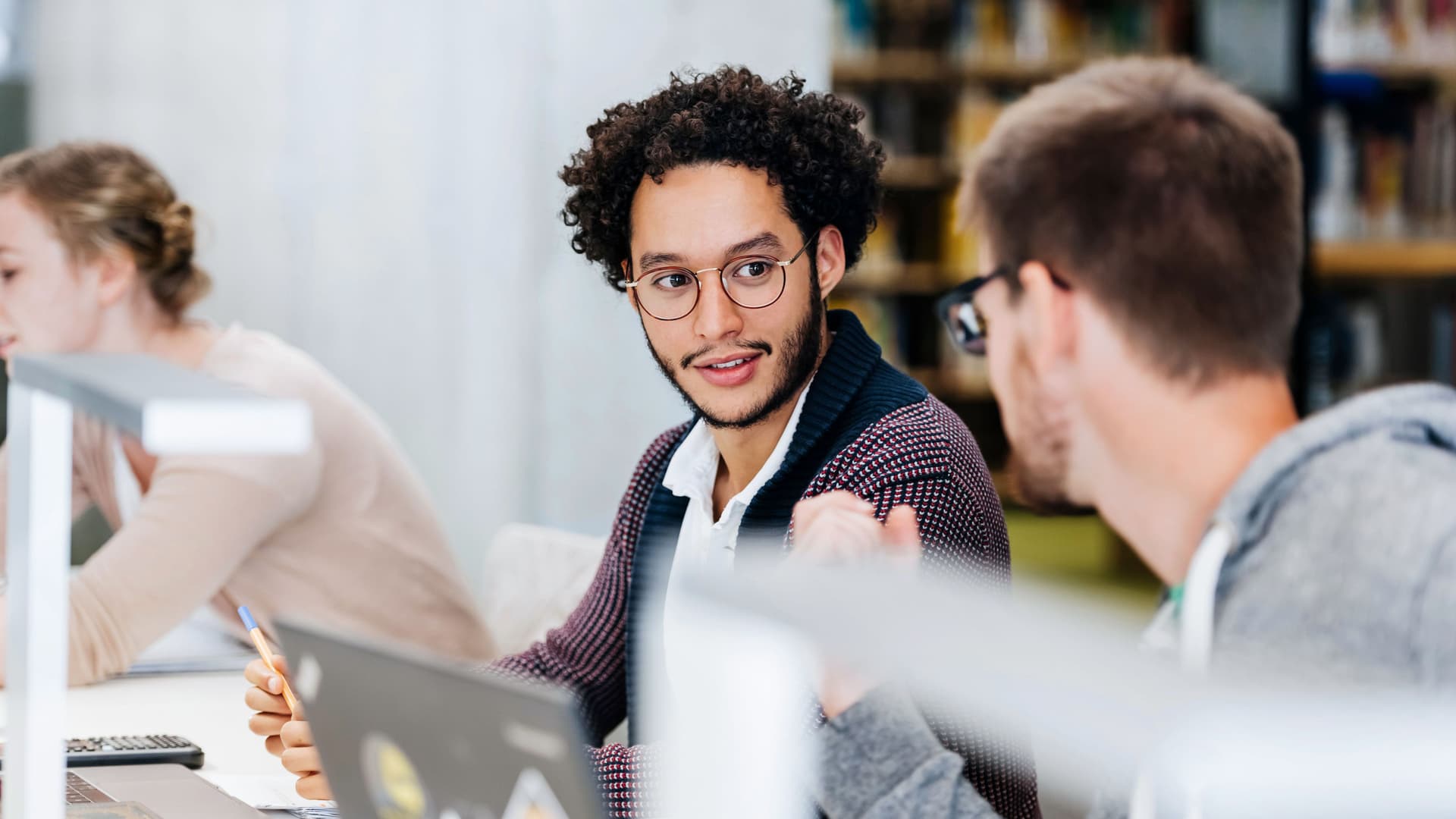 Zwei Studenten vor ihren Laptops, die sich beim Lernen in der Bibliothek leise unterhalten.