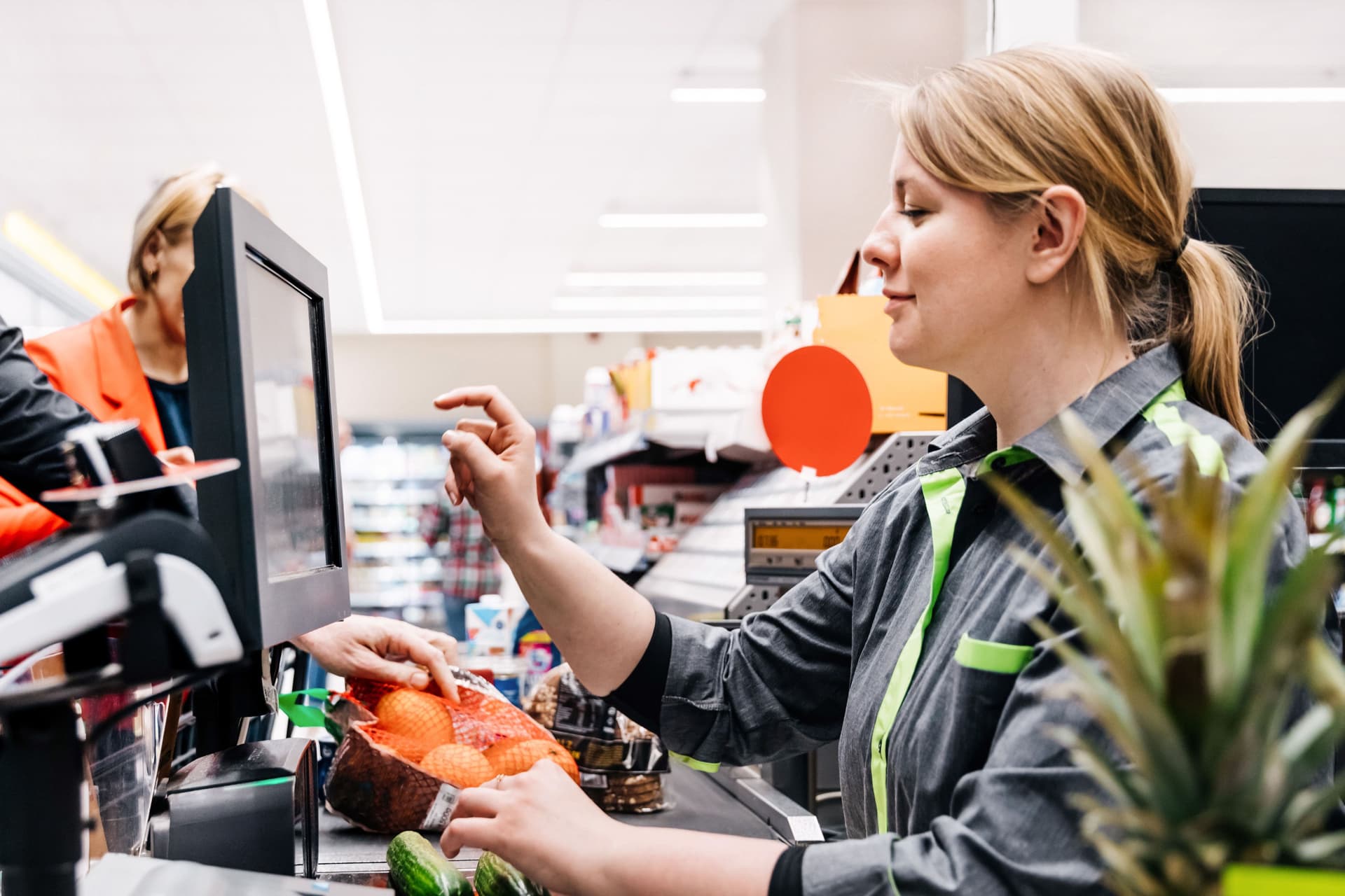 Ein Supermarktkassiererin tippt auf einen Bildschirm während zwei Gurken auf dem Scanner liegen. Ein männlicher Kunde legt derweil Waren auf das Band.