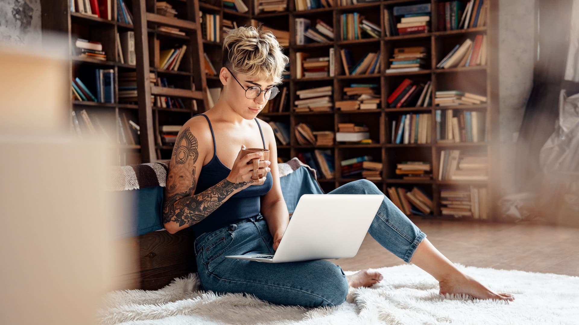 Junge Frau mit kurzen Haaren und Brille, die entspannt mit einem Laptop auf dem Schoß auf dem Boden sitzt und sich etwas ansieht. Sie hat eine Tasse in der rechten Hand.