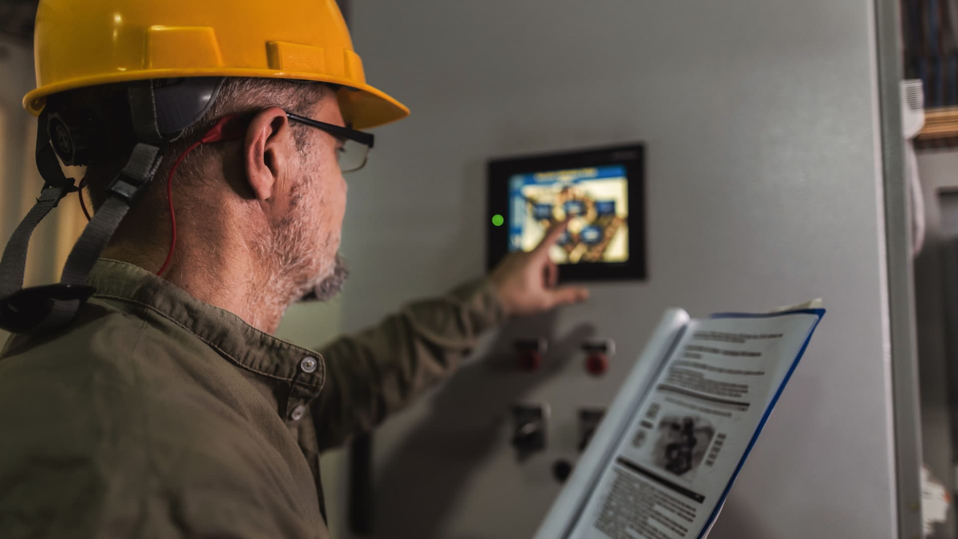Mann mit gelbem Helm bedient ein Display an der Wand und hält eine Bedienungsanleitung in der Hand. 