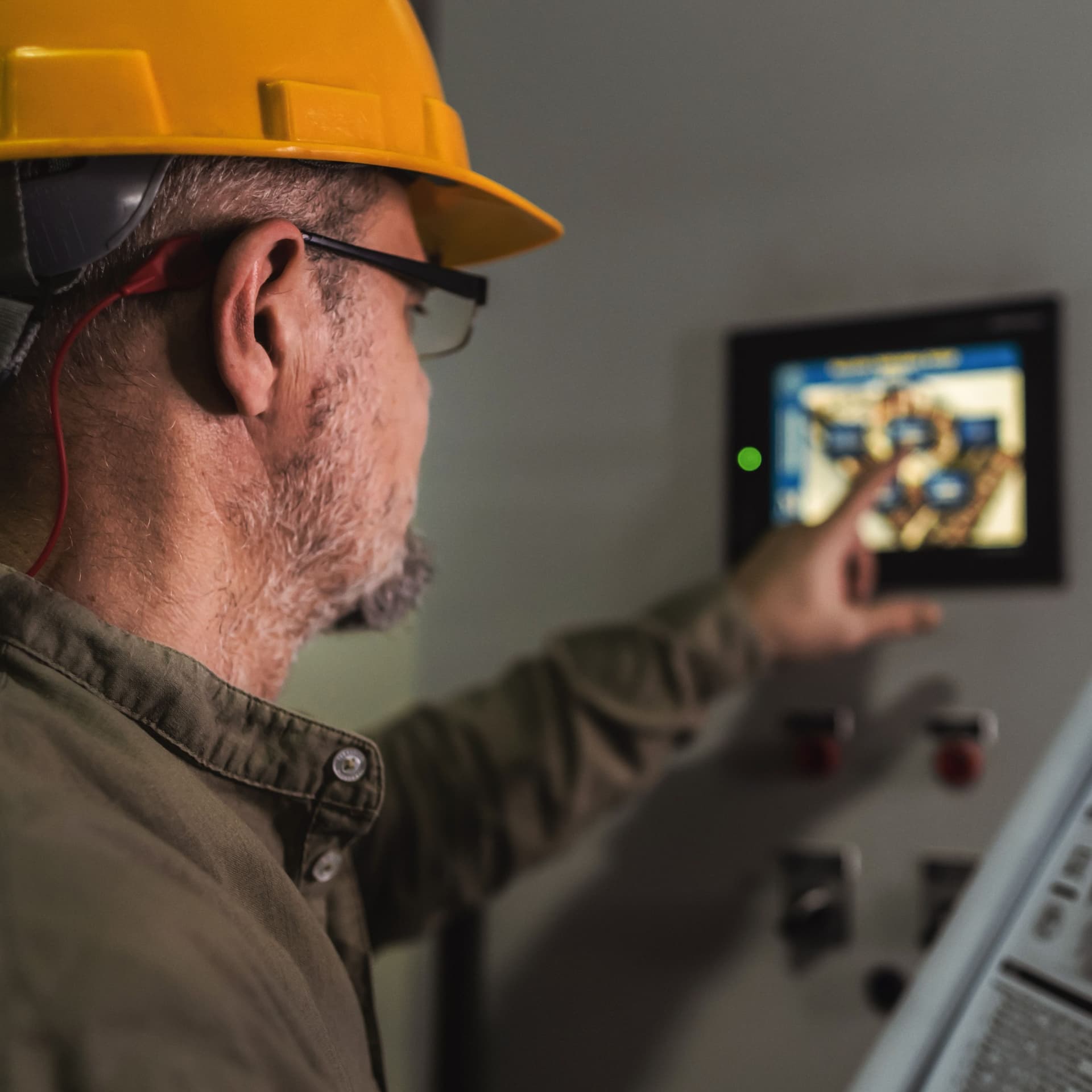 Mann mit gelbem Helm bedient ein Display an der Wand und hält eine Bedienungsanleitung in der Hand. 