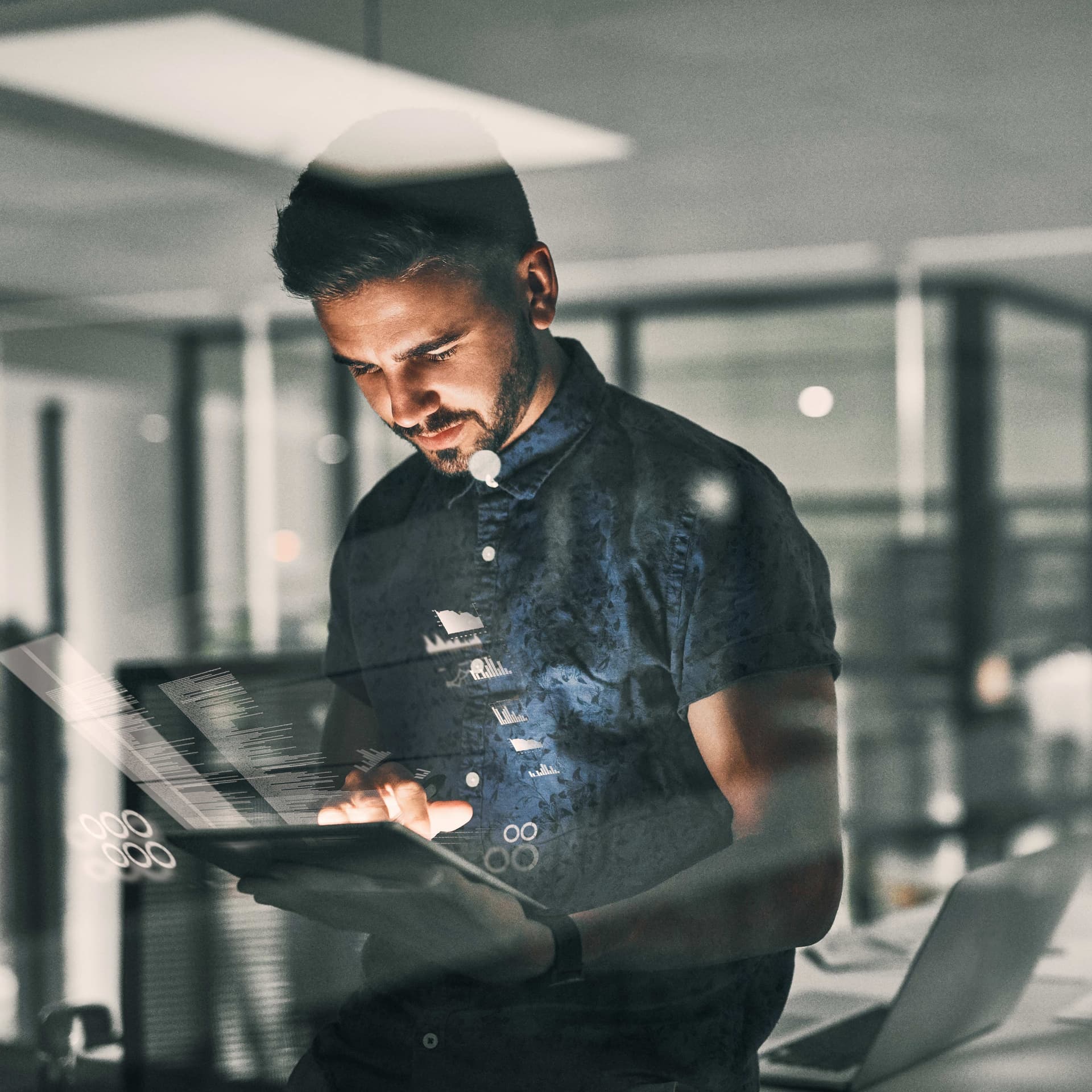 Junger Mann schaut im halbdunklen auf ein Tablet. Das Büro hinter ihm ist leer.