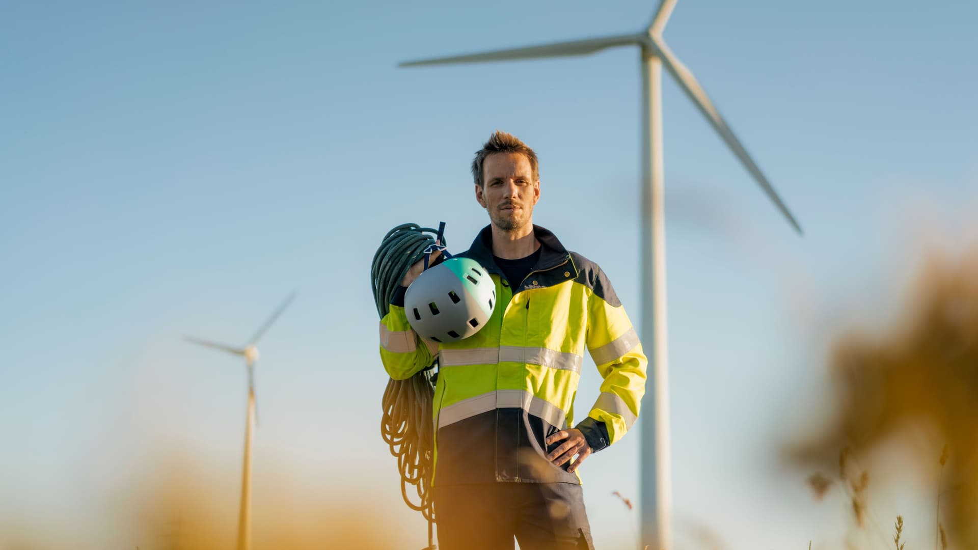 Techniker schaut in die Kamera. Er steht auf einem Feld in einem Windpark mit Kletterausrüstung über der Schulter.