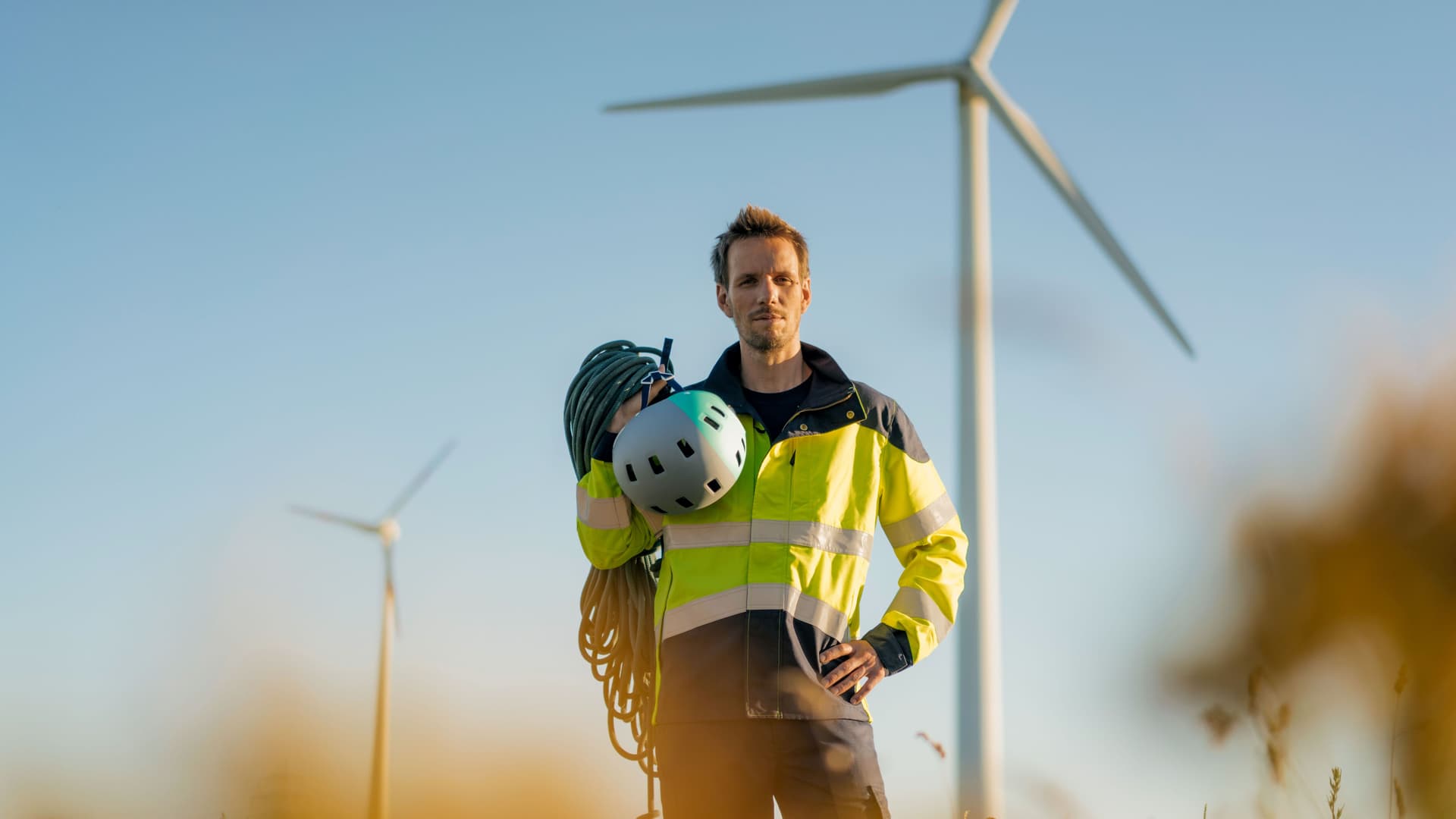 Techniker schaut in die Kamera. Er steht auf einem Feld in einem Windpark mit Kletterausrüstung über der Schulter.