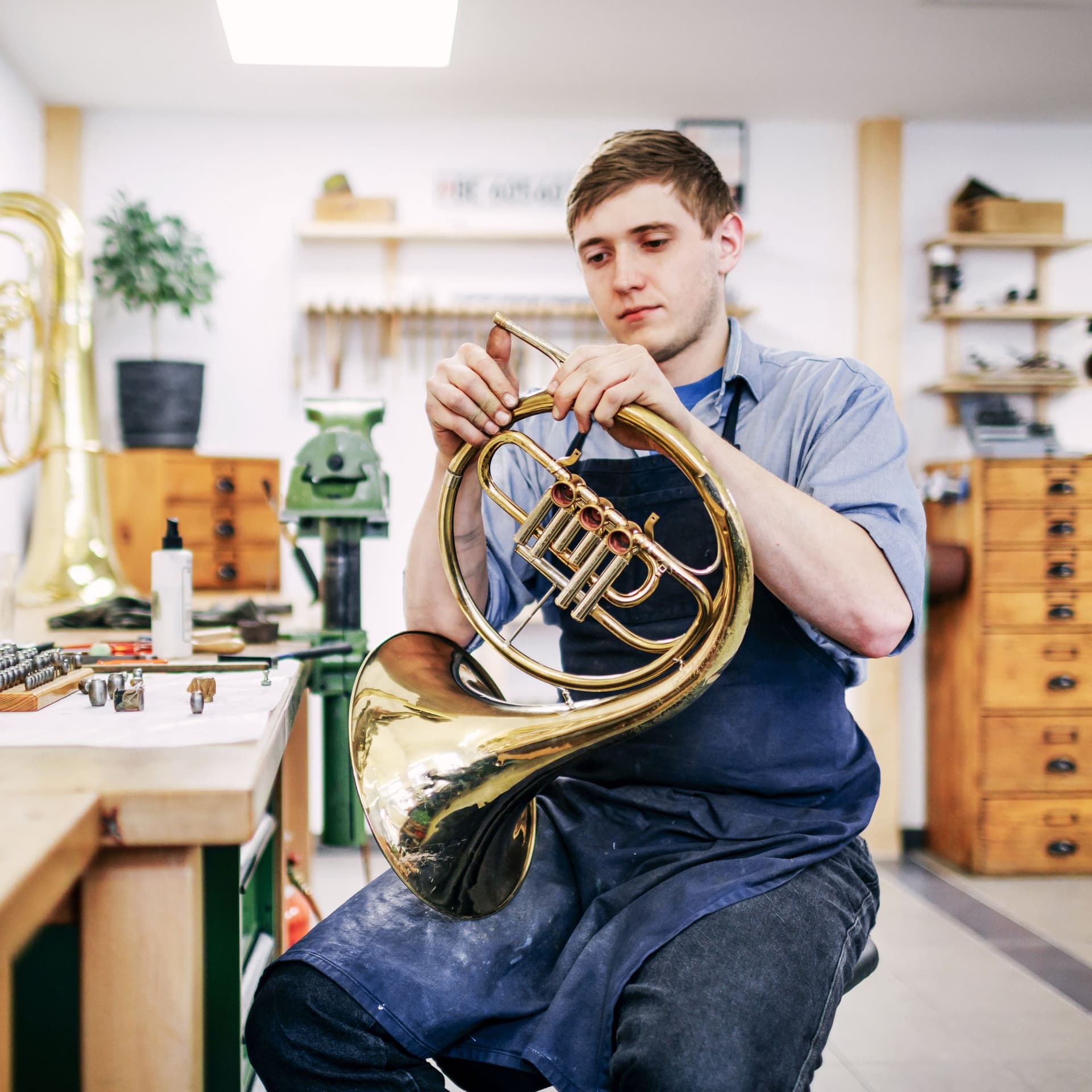 Ein junger Mann sitzt in einer Werkstatt und arbeitet an einem Blasinstrument. Er trägt eine Schürze und ist ganz vertieft.