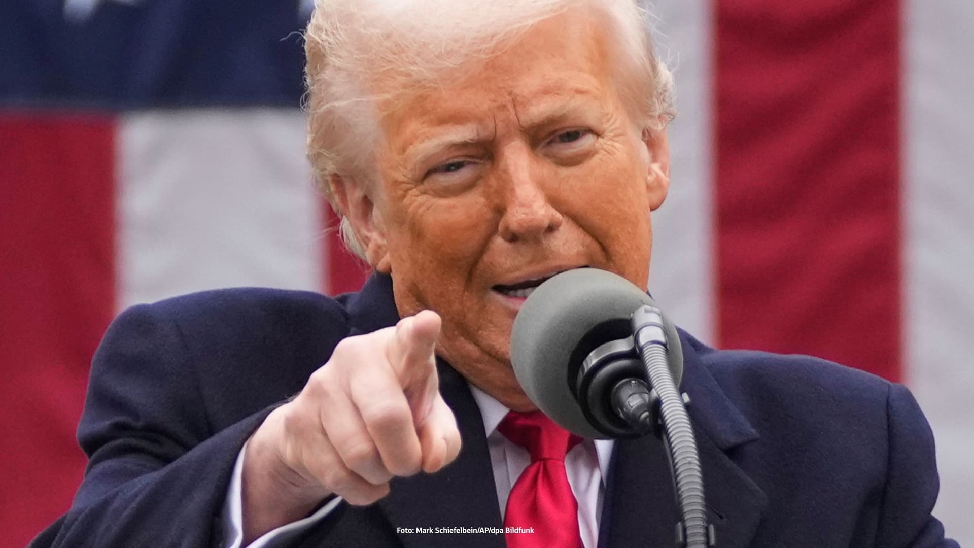 Donald Trump spricht in ein Mikrofon und zeigt mit dem Finger nach vorne. Im Hintergrund ist eine große US-Flagge zu sehen. 