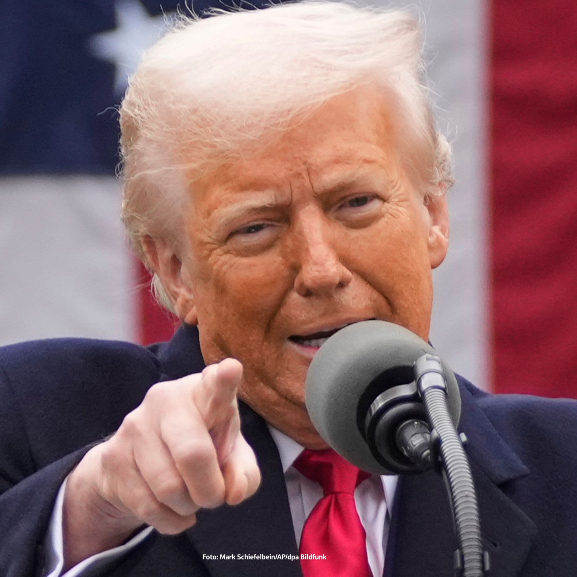 Donald Trump spricht in ein Mikrofon und zeigt mit dem Finger nach vorne. Im Hintergrund ist eine große US-Flagge zu sehen. 