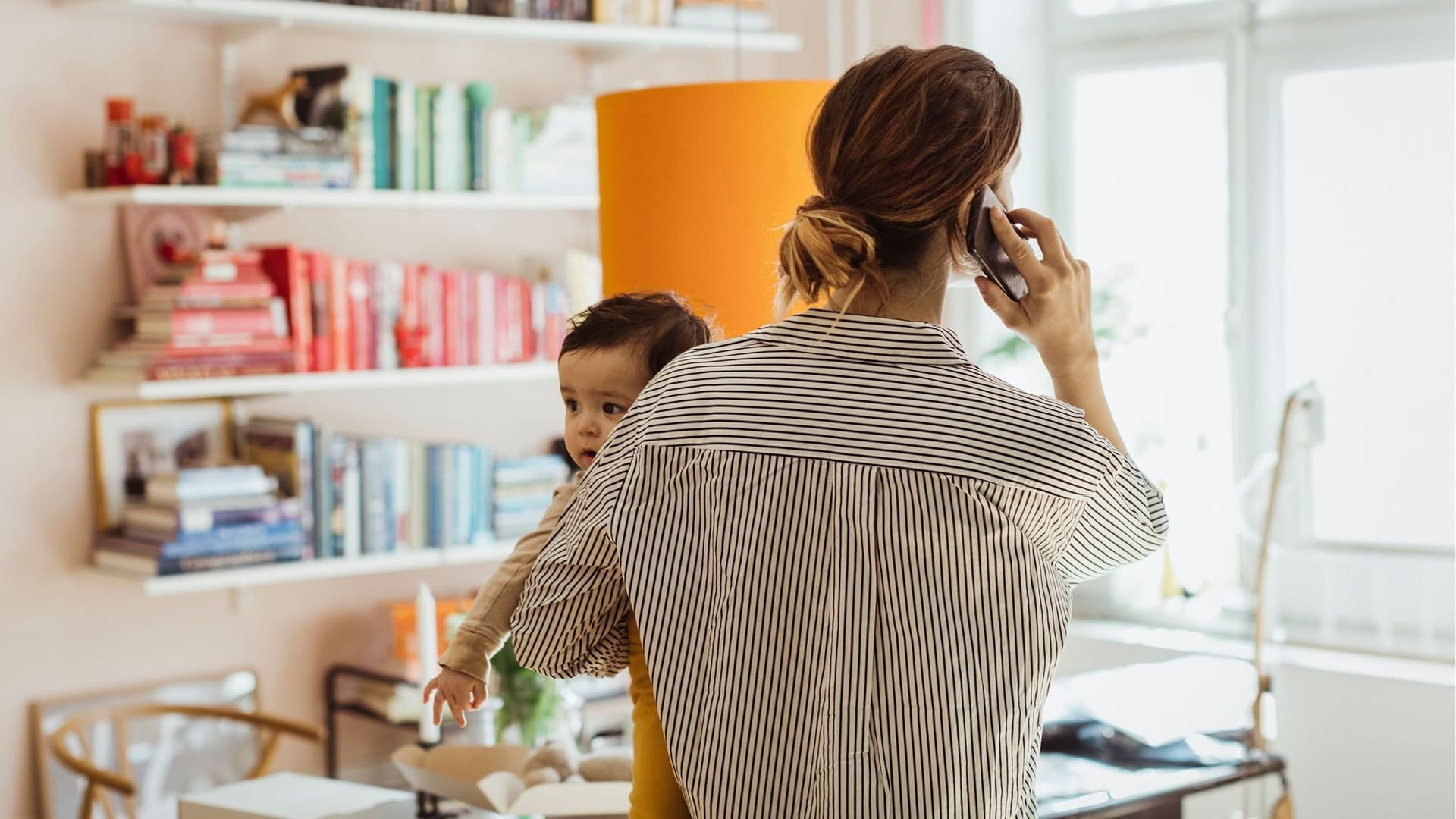 Eine junge Mutter hält zuhause, vor ihrem Schreibtisch telefonierend, ihr Baby auf dem Arm und blickt aus dem Fenster.