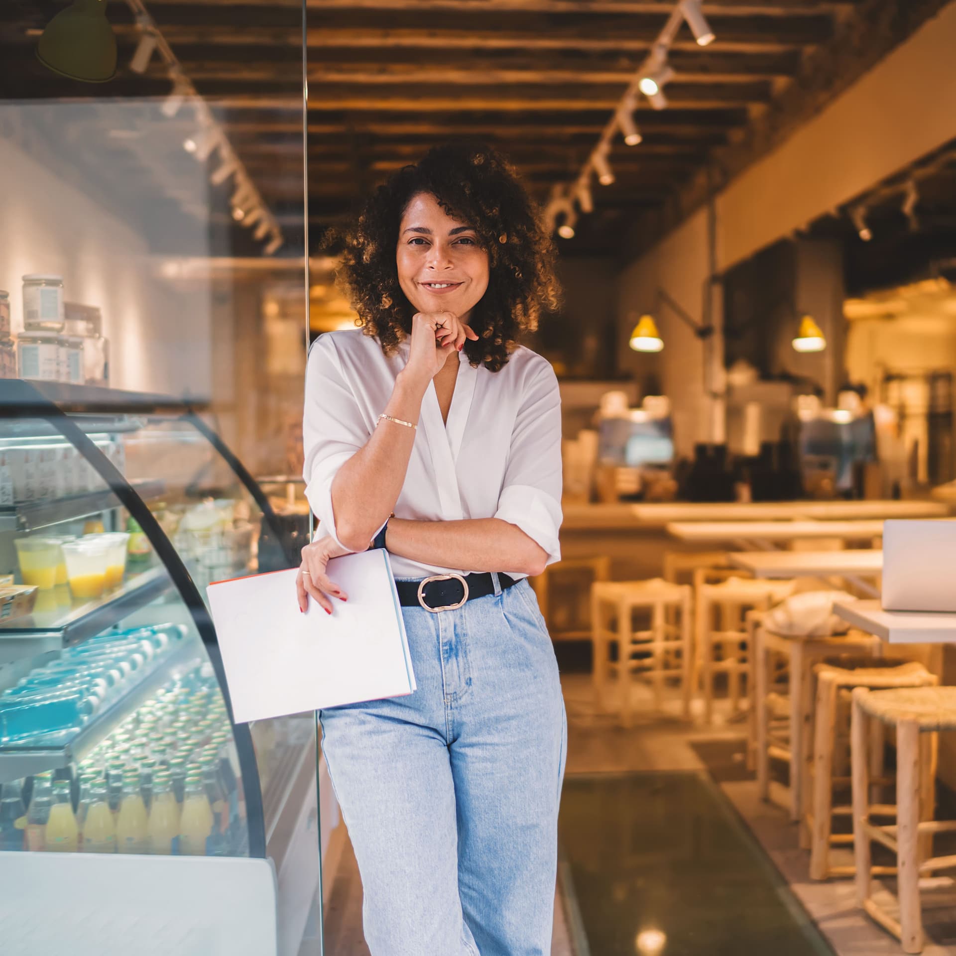 Junge Frau mit Locken steht lächeln am Eingang eines Cafes. Sie hat Unterlagen in der Hand und lehnt sich an die Glastür.