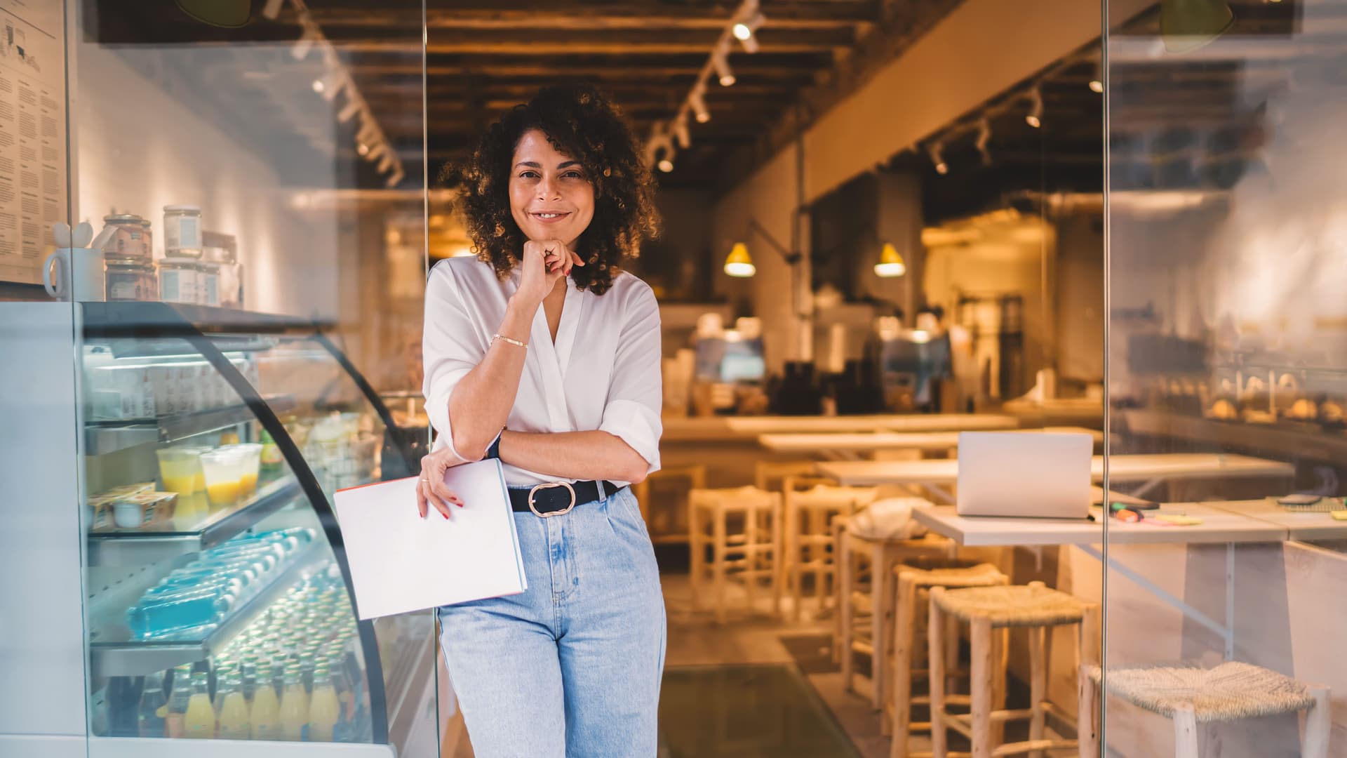 Junge Frau mit Locken steht lächeln am Eingang eines Cafes. Sie hat Unterlagen in der Hand und lehnt sich an die Glastür.