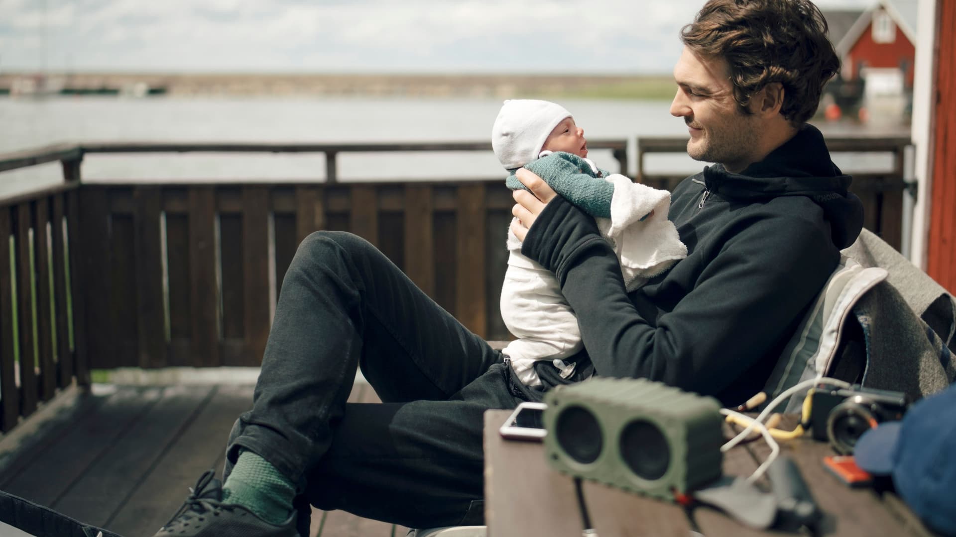 Junger Vater sitzt auf einer Bank an der Küste und lächelt sein Baby an.