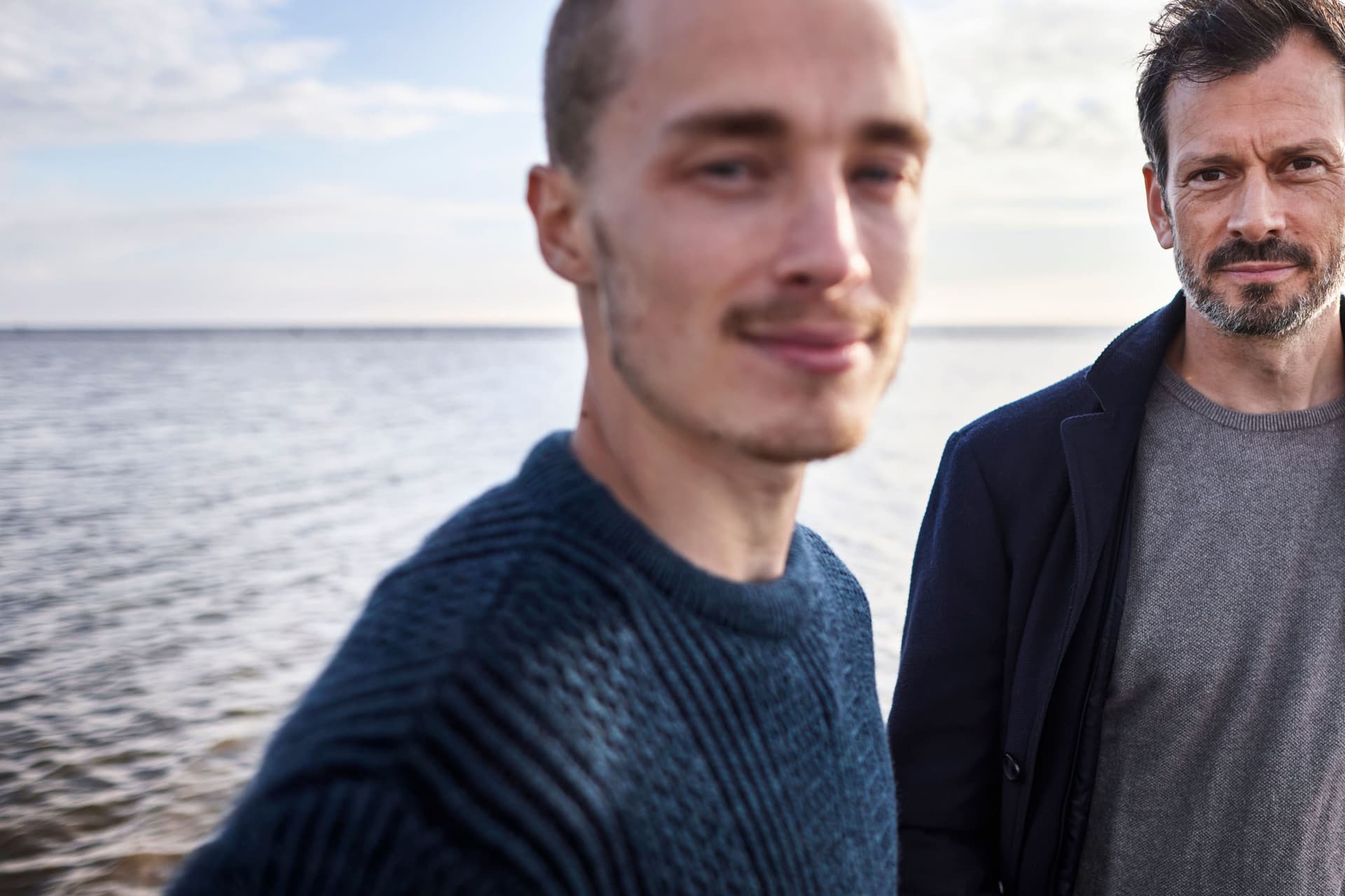 Porträt von einem älteren und einem jüngeren Mann am Meer bei bewölktem Himmel.