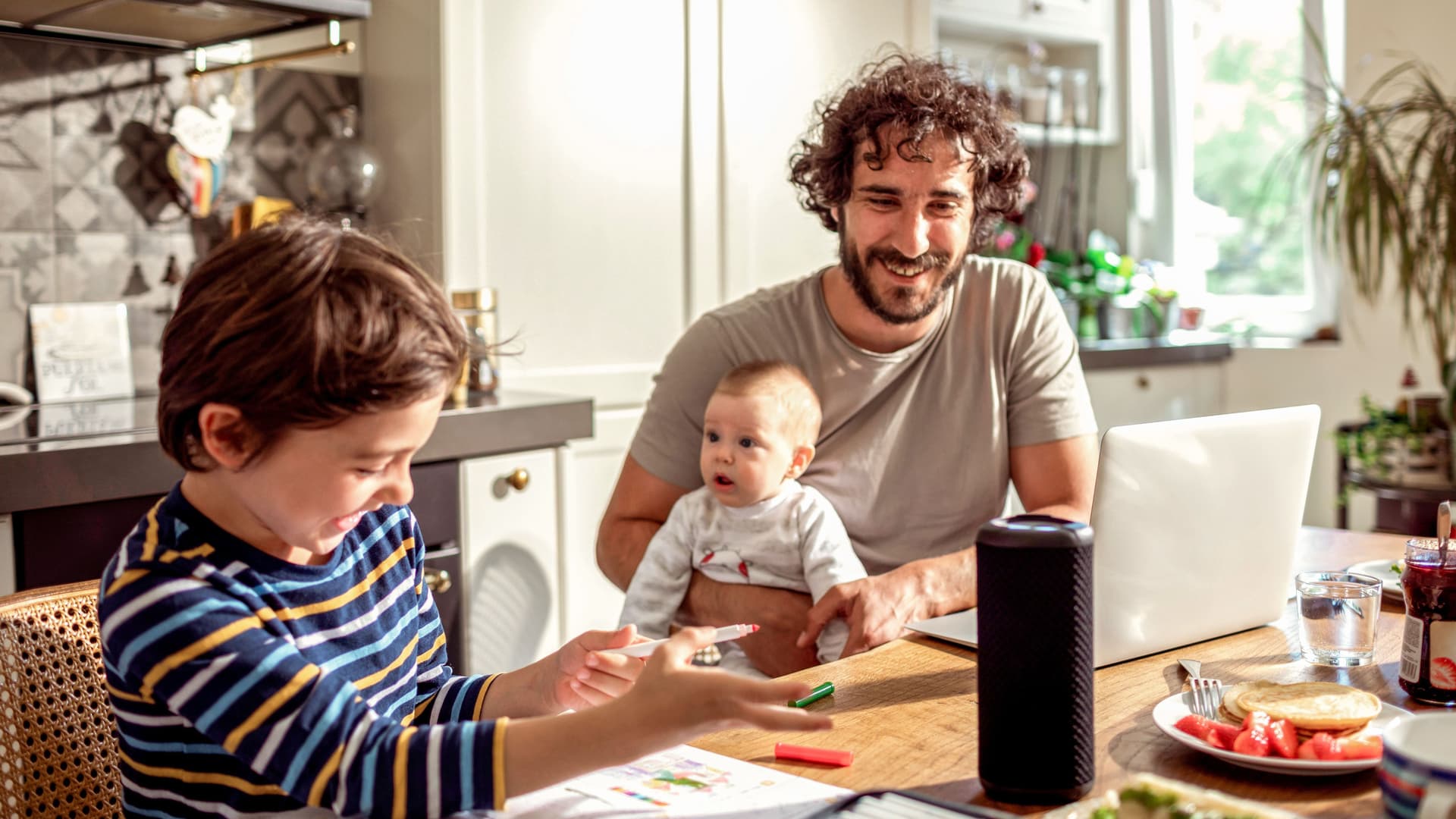 Vater mit Baby auf dem Schoß sitzen am Küchentisch vor einem Smart Speaker. Neben ihm sitzt ein Junge im Grundschulalter, der seine Hausaufgaben löst. 