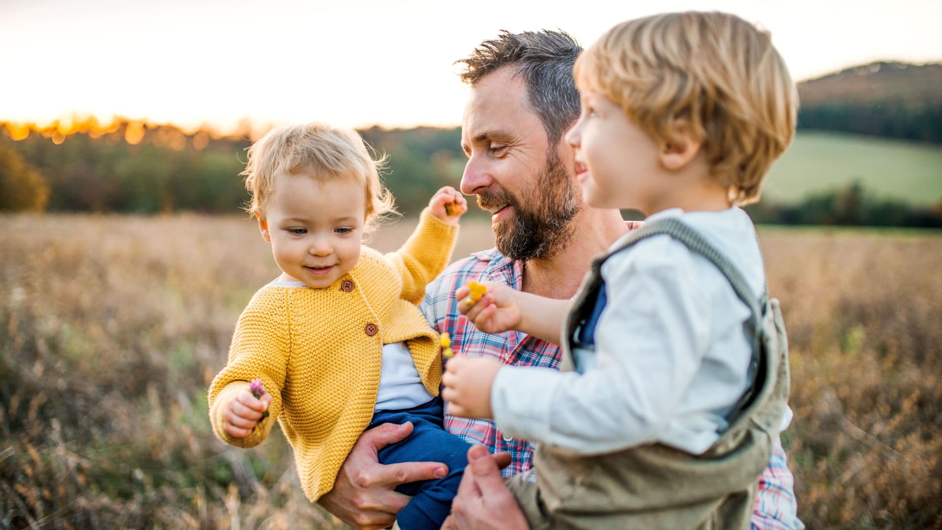 Ein Vater mit zwei Kleinkindern auf dem Arm auf einer Herbstwiese im Freien.