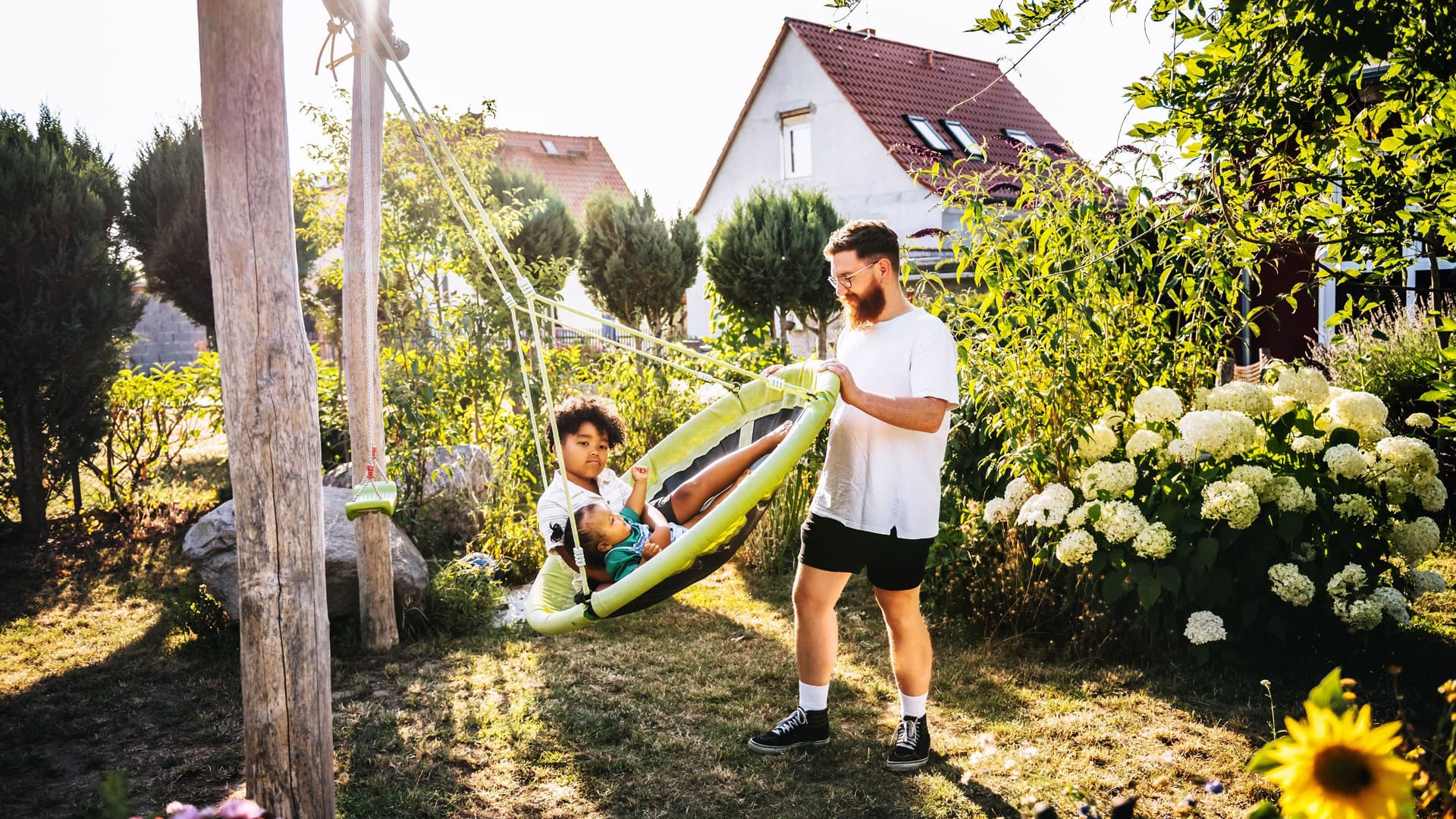 Ein Mann schubst eine große Schaukel mit zwei Kindern in einem grünen Garten an. Im Hintergrund sieht man Einfamilienhäuser.