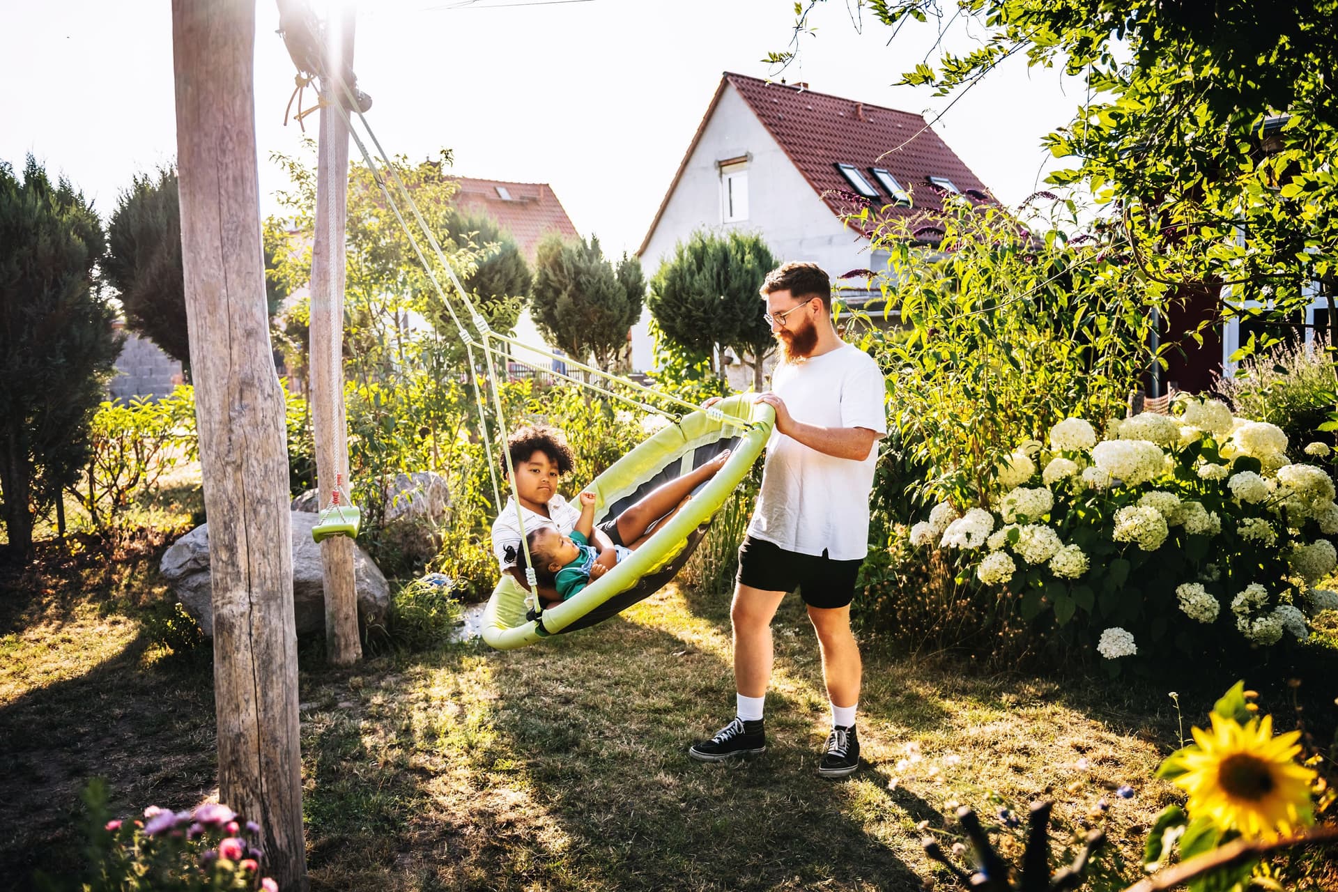 Ein Mann schubst eine große Schaukel mit zwei Kindern in einem grünen Garten an. Im Hintergrund sieht man Einfamilienhäuser.