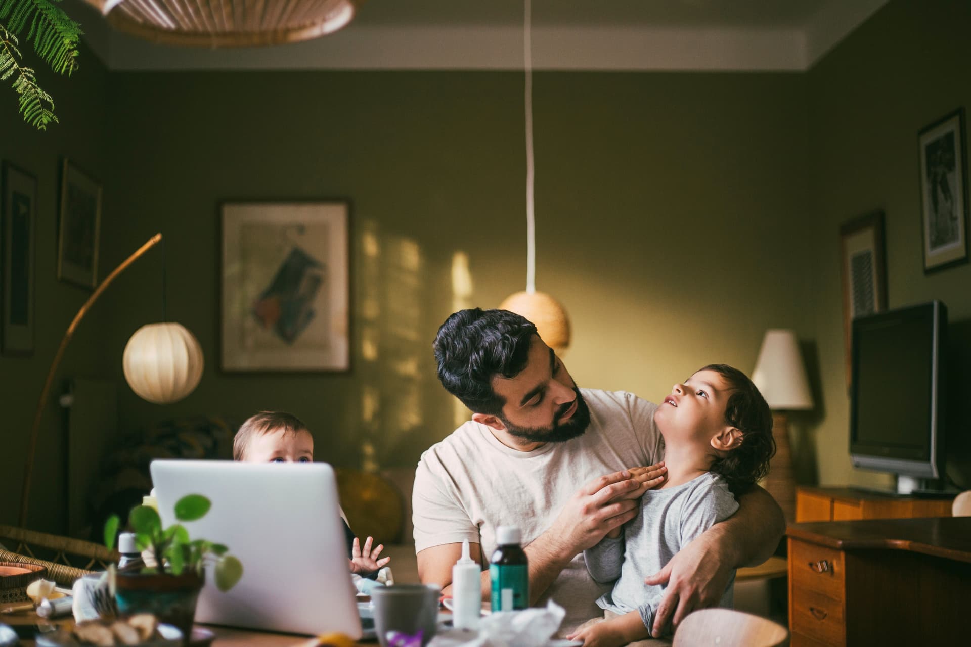 Ein Vater sitzt zuhause mit zwei Kindern vor einem Laptop. Neben dem Laptop stehen Medikamente und der Vater untersucht den Hals seines Sohnes.