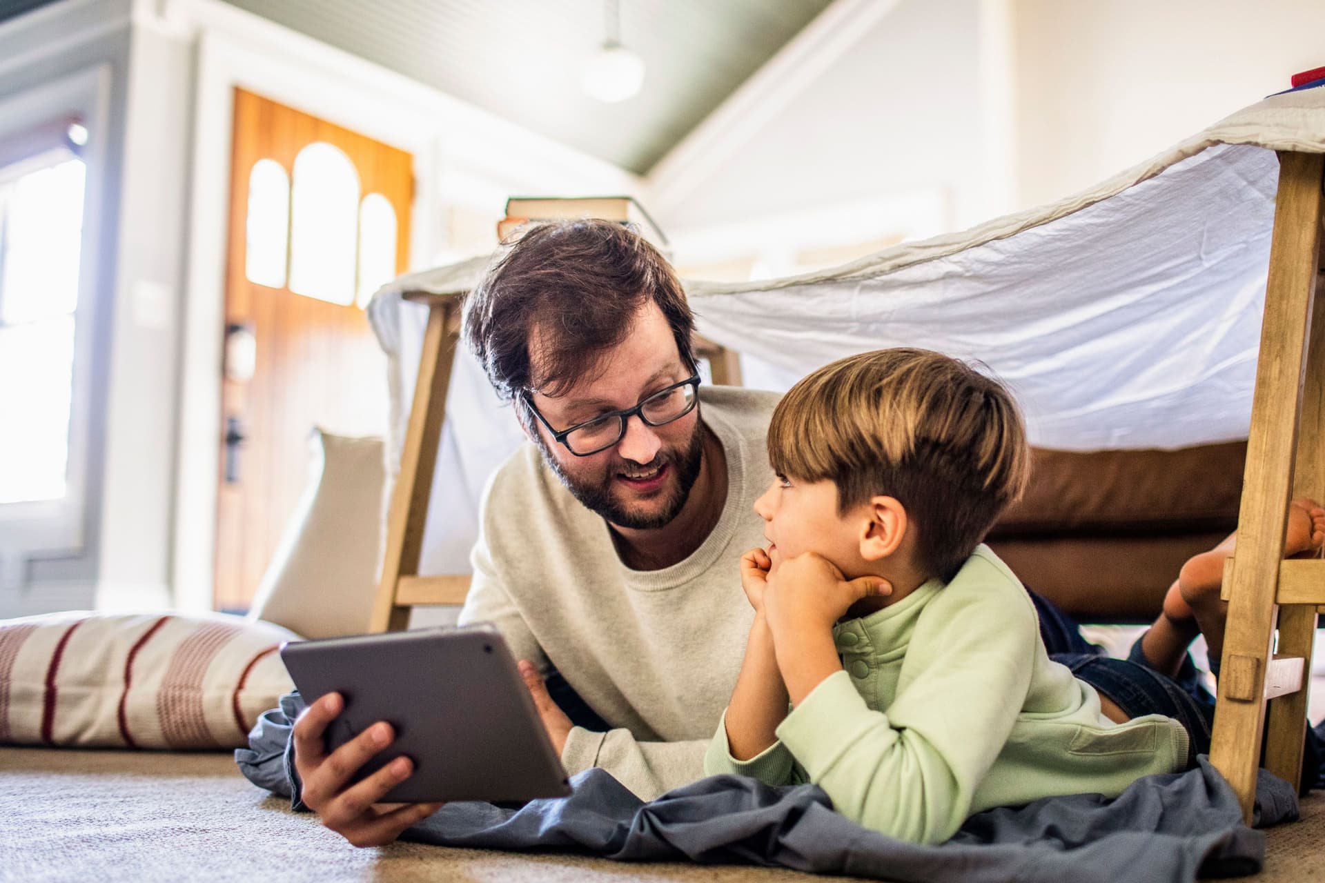 Mann und Kind liegen zusammen mit einem Tablet auf dem Boden. Über ihnen ist ein Bettlaken über zwei Stühlen gespannt.