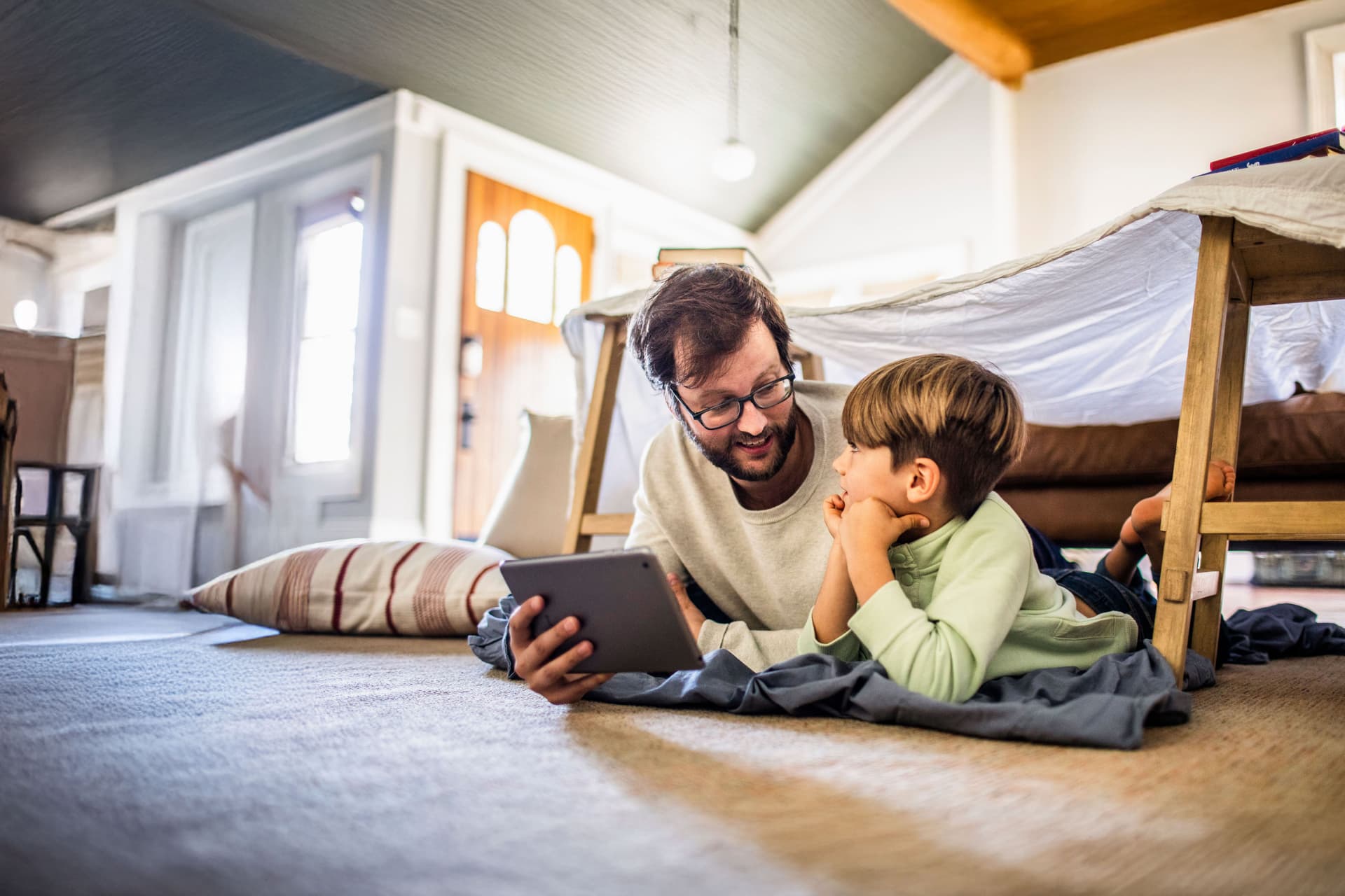 Mann und Kind liegen zusammen mit einem Tablet auf dem Boden. Über ihnen ist ein Bettlaken über zwei Stühlen gespannt.