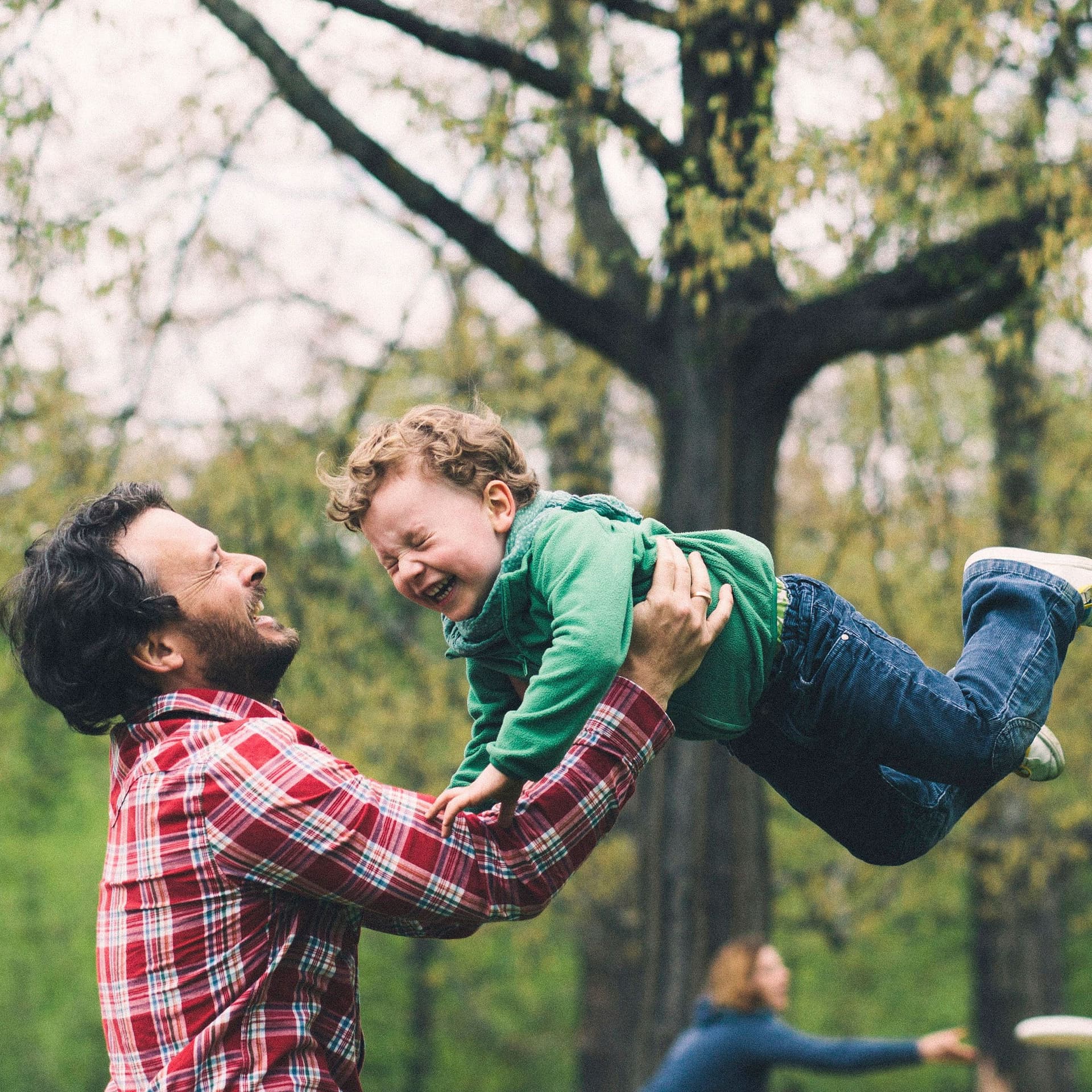 Vater wirbelt seinen Sohn im Park herum. Die Mutter spielt im Hintergrund Frisbee.