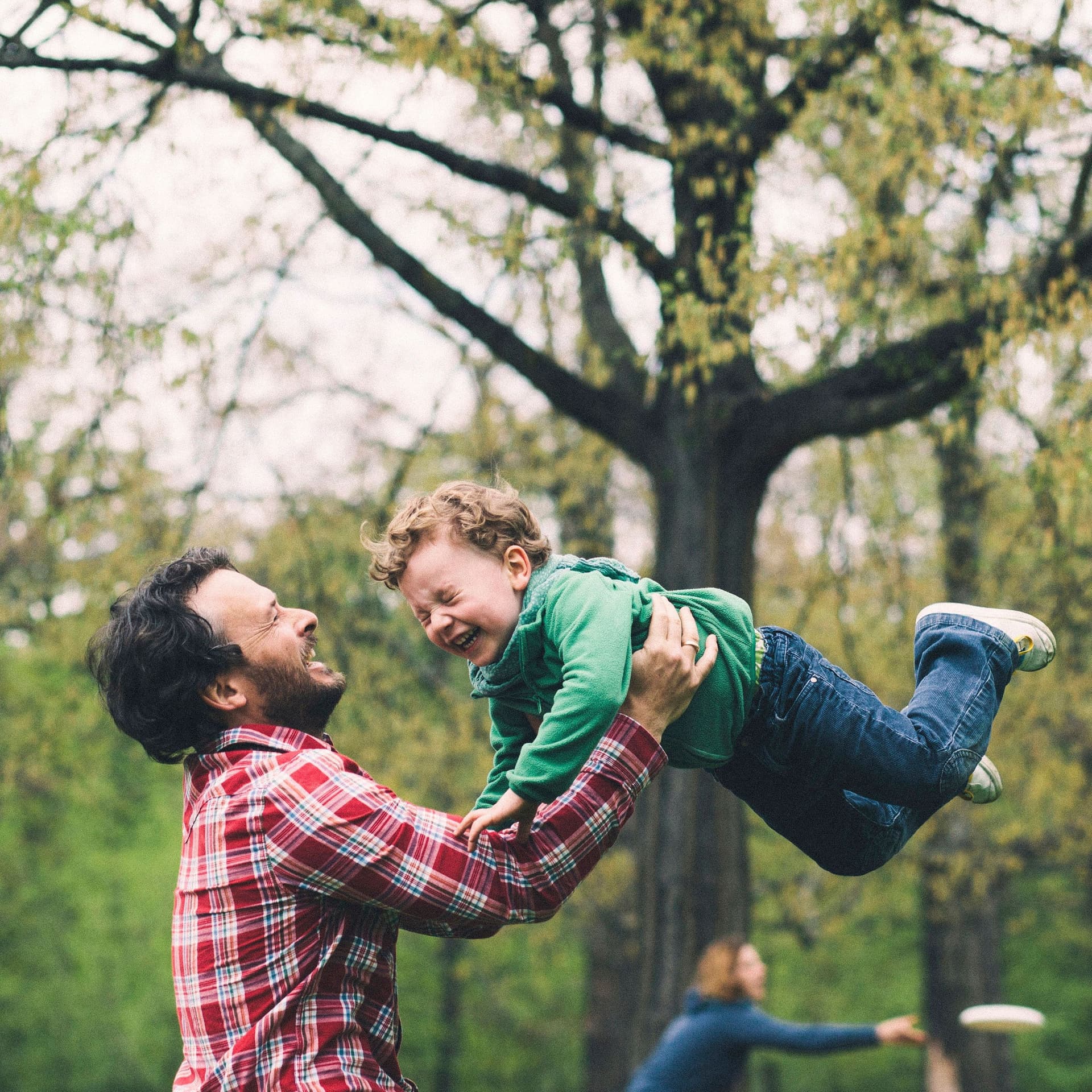 Vater wirbelt seinen Sohn im Park herum. Die Mutter spielt im Hintergrund Frisbee.